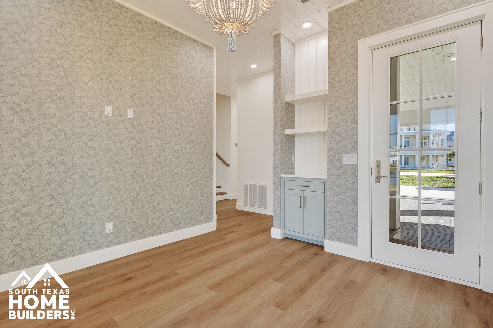 Bright empty foyer with wood floors, patterned wallpaper, white trim, built-in shelving, and glass front door