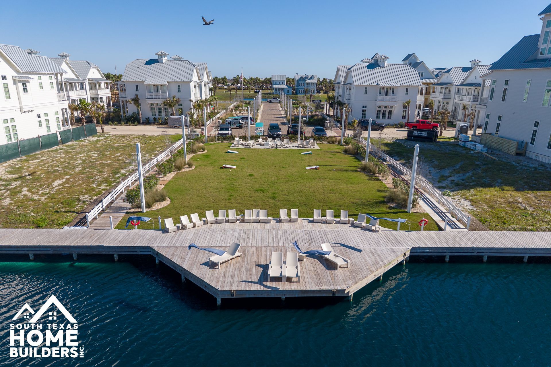 Waterfront view of white homes around a landscaped dock and lawn, with a “Home Builders” logo in the corner