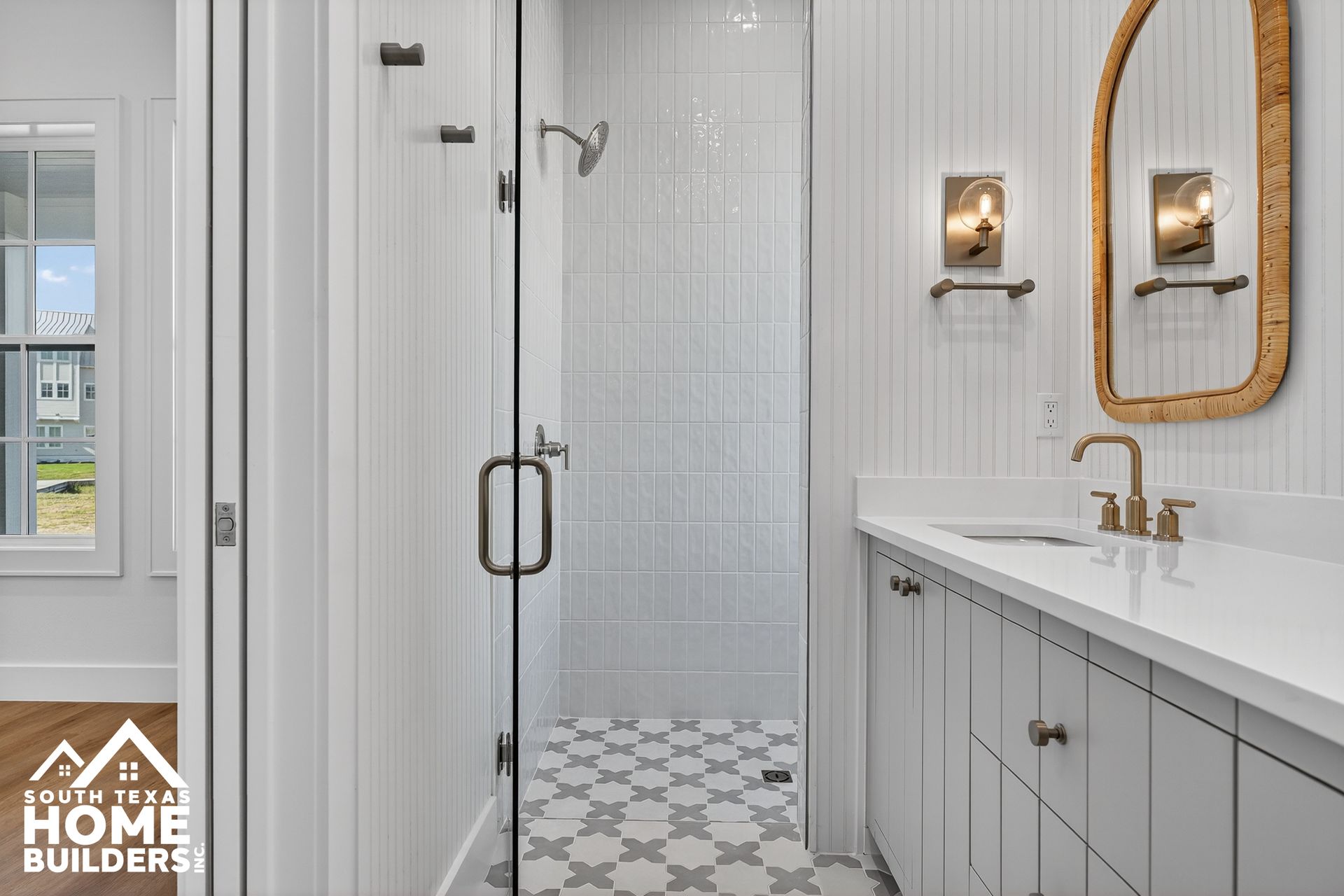 Bright white bathroom with glass shower, patterned tile floor, and vanity with gold accents.
