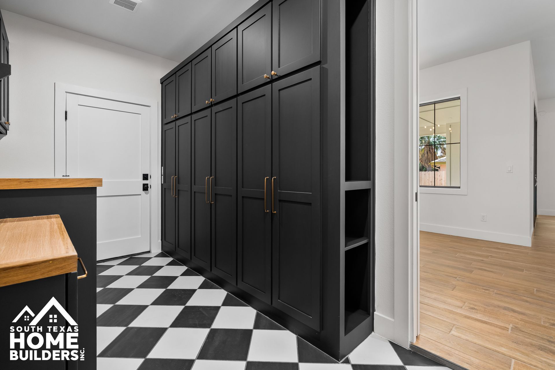 Black cabinetry in a hallway with checkered floor. A white door is on the left; a room with wood flooring is to the right.