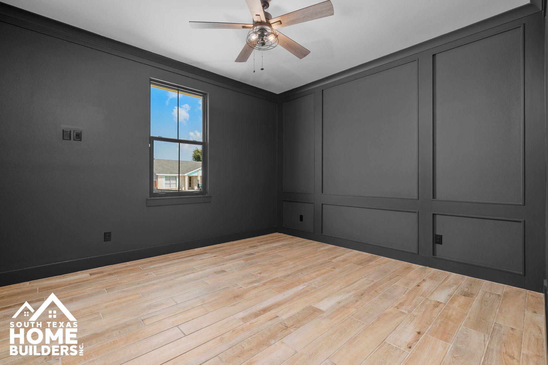 Dark gray paneled room with light wood floor, window, and ceiling fan.