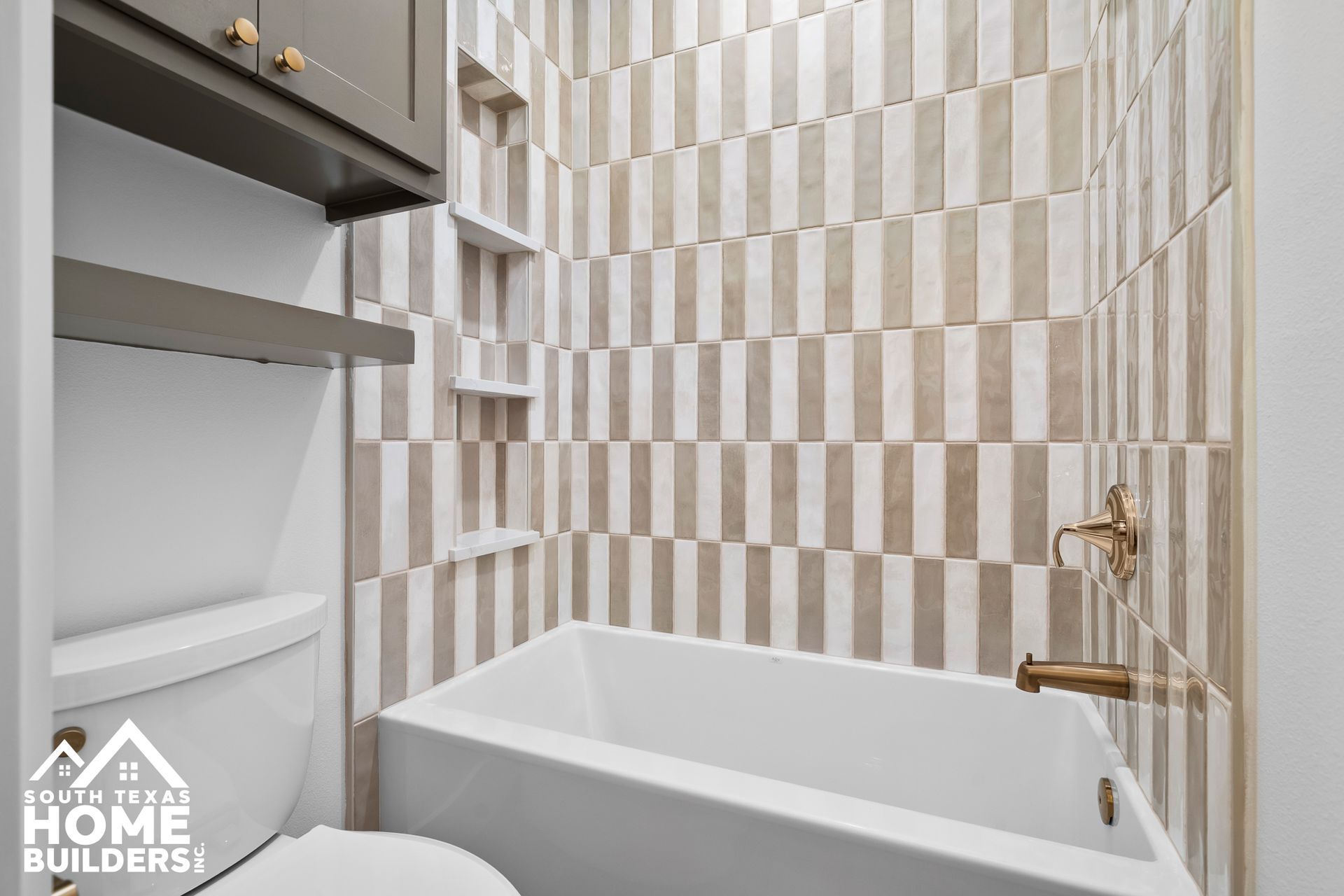 Small bathroom with white tub, vertical beige and white tile, and gold fixtures.