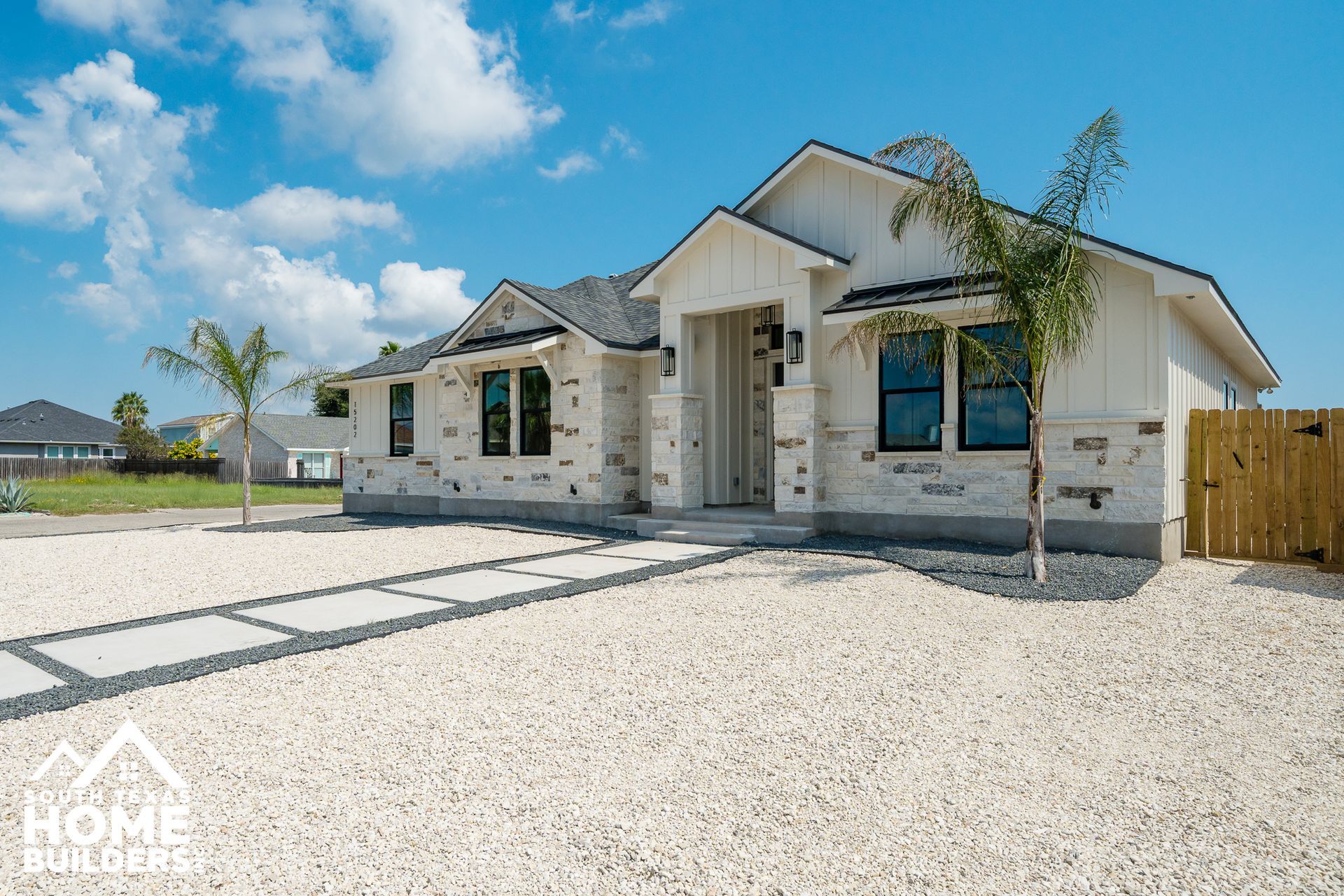 White-sided house with stone accents, palm trees, and gravel driveway under a bright blue sky.