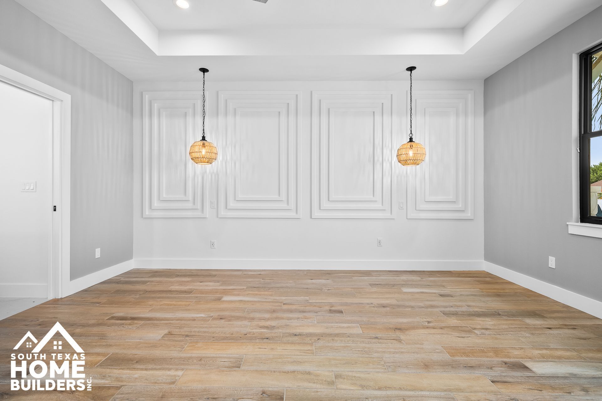 Empty room with light wood floor, white walls with rectangular paneling, and two pendant lights.