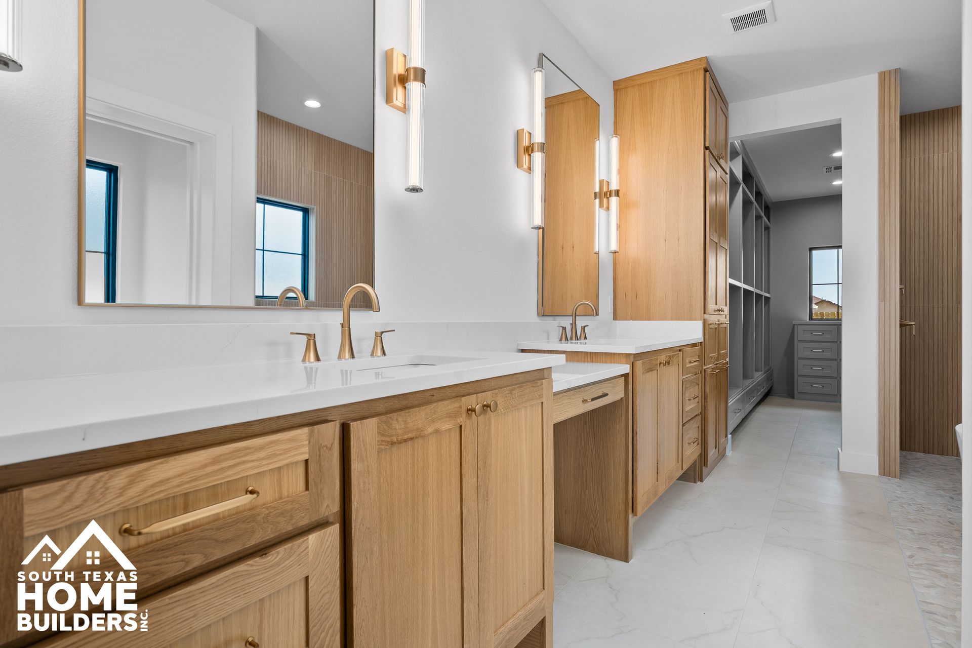 Modern bathroom with light wood cabinets, white countertops, gold fixtures, and a walk-in closet.