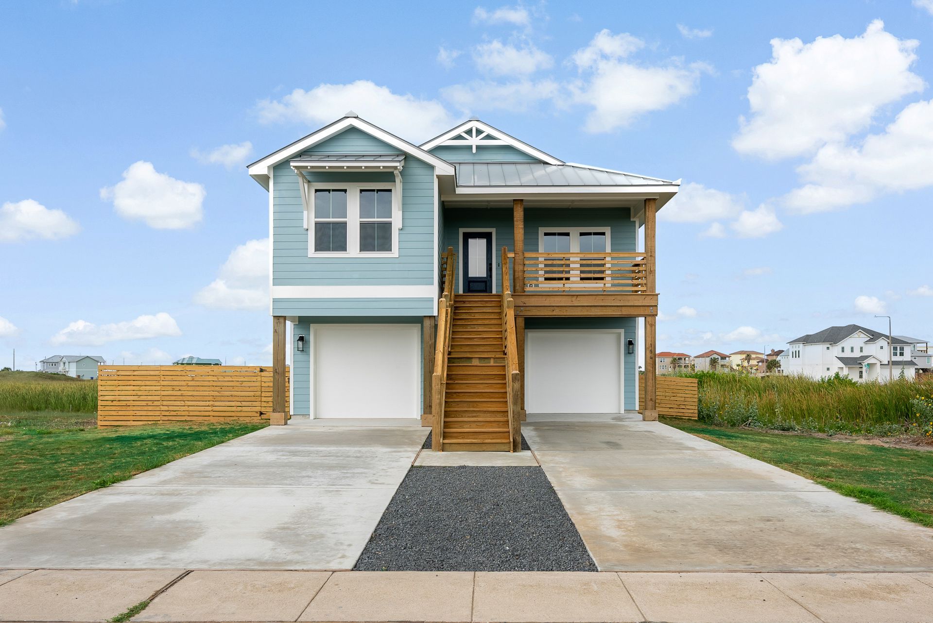 Two-story light blue house with two garages, a wooden staircase, and a porch under a blue sky.