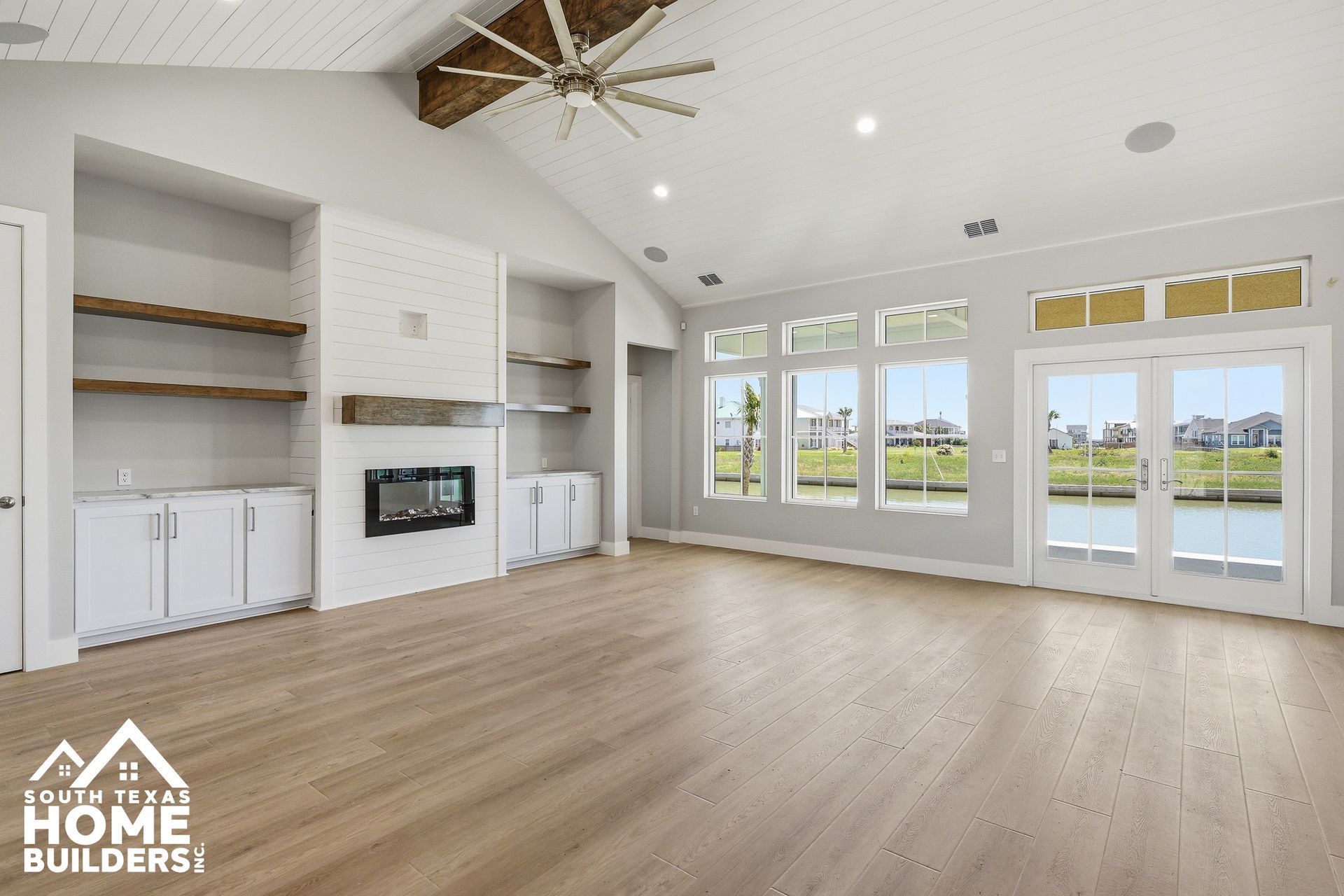 Bright empty living room with wood floors, white built-in shelves, fireplace, and large windows overlooking grass.