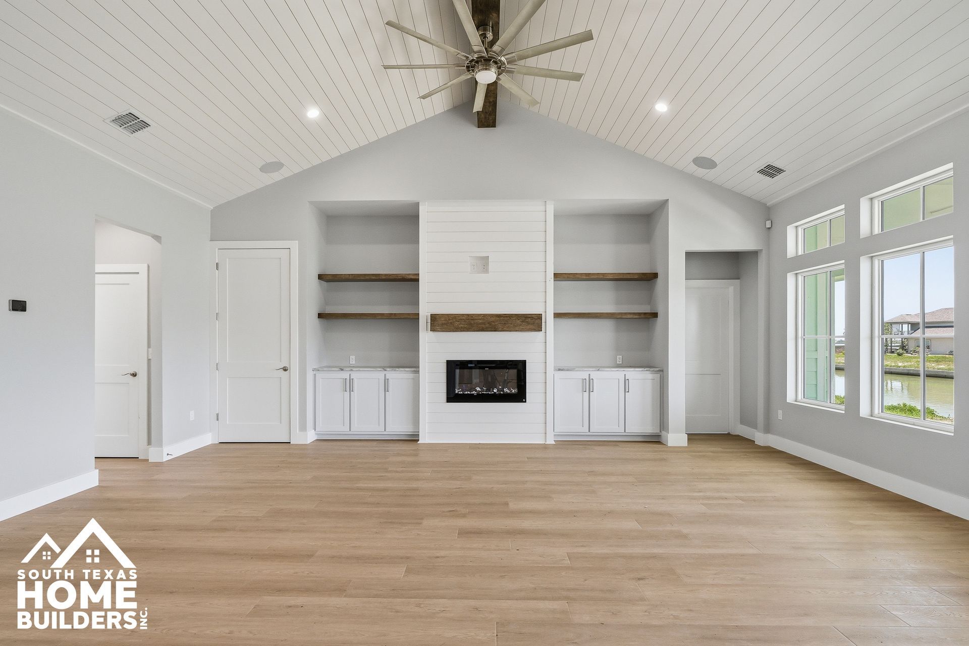 Bright vaulted living room with fireplace, built-in shelves, light wood floors, and large windows