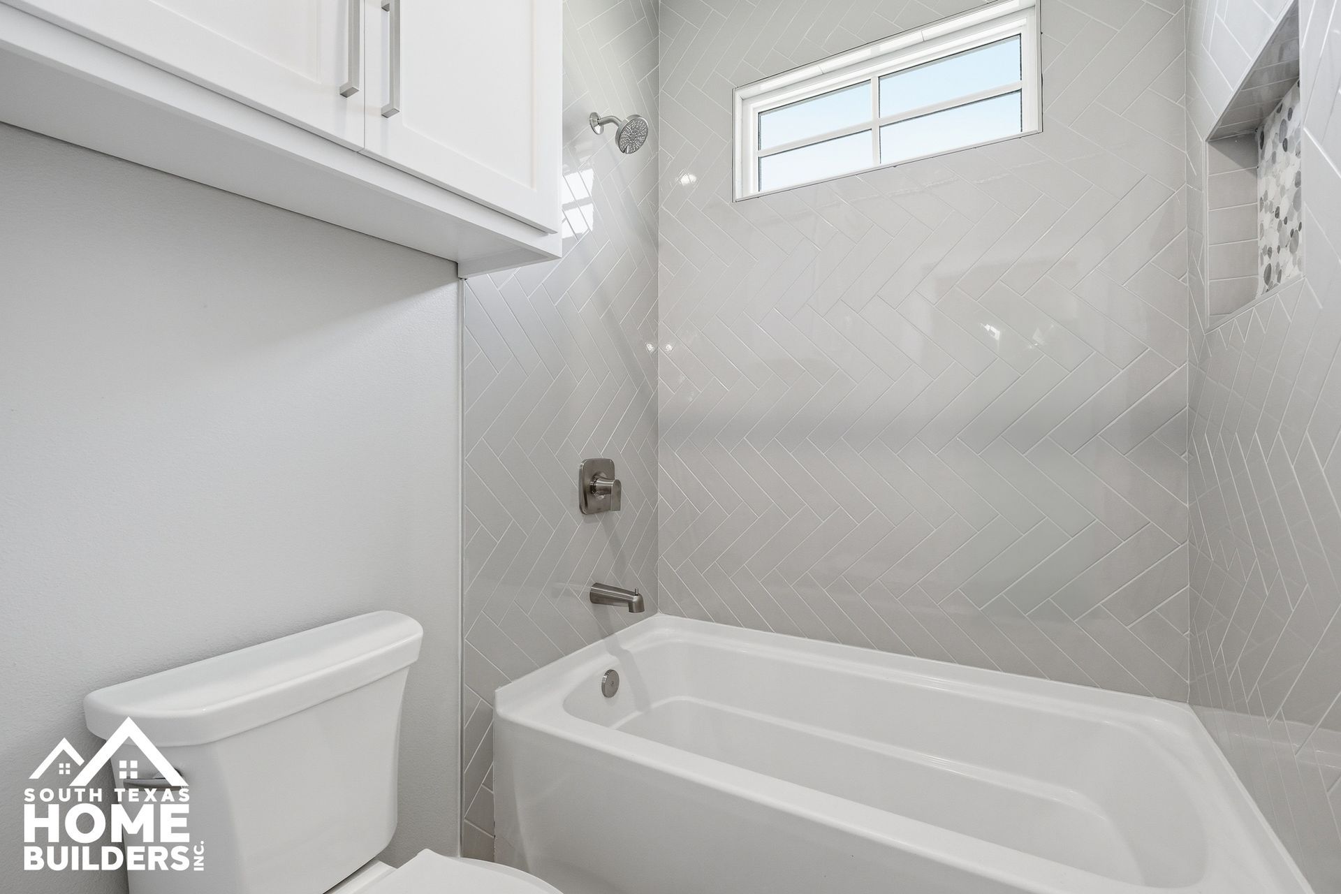 Bright white bathroom with a tub, showerhead, window, and cabinet above the toilet.