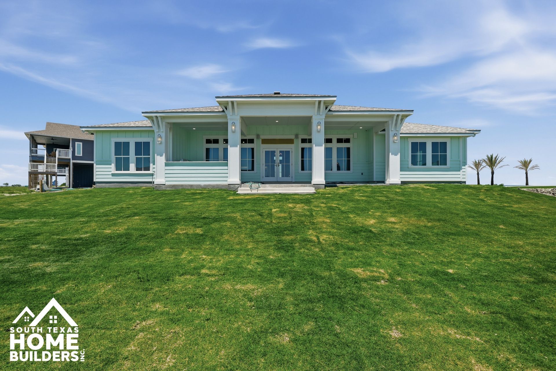 Modern mint-green beachfront house with white trim and a large grassy lawn under a blue sky