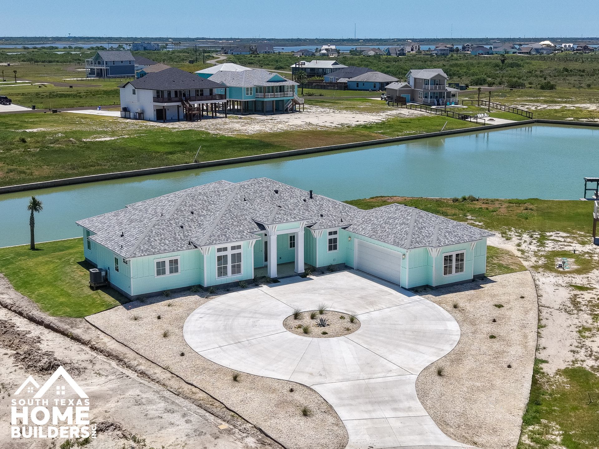Mint-green waterfront house with white roof and circular driveway in a grassy residential area