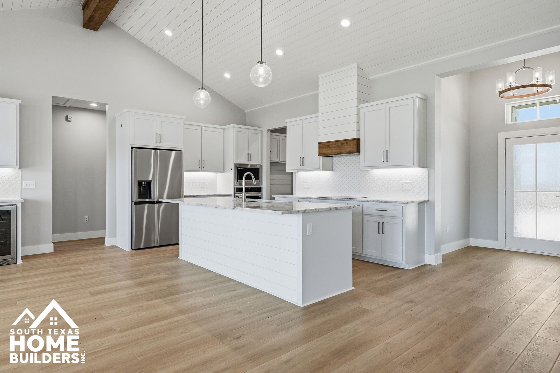 Bright white open-plan kitchen and living area with island, stainless fridge, vaulted ceiling, and wood floors