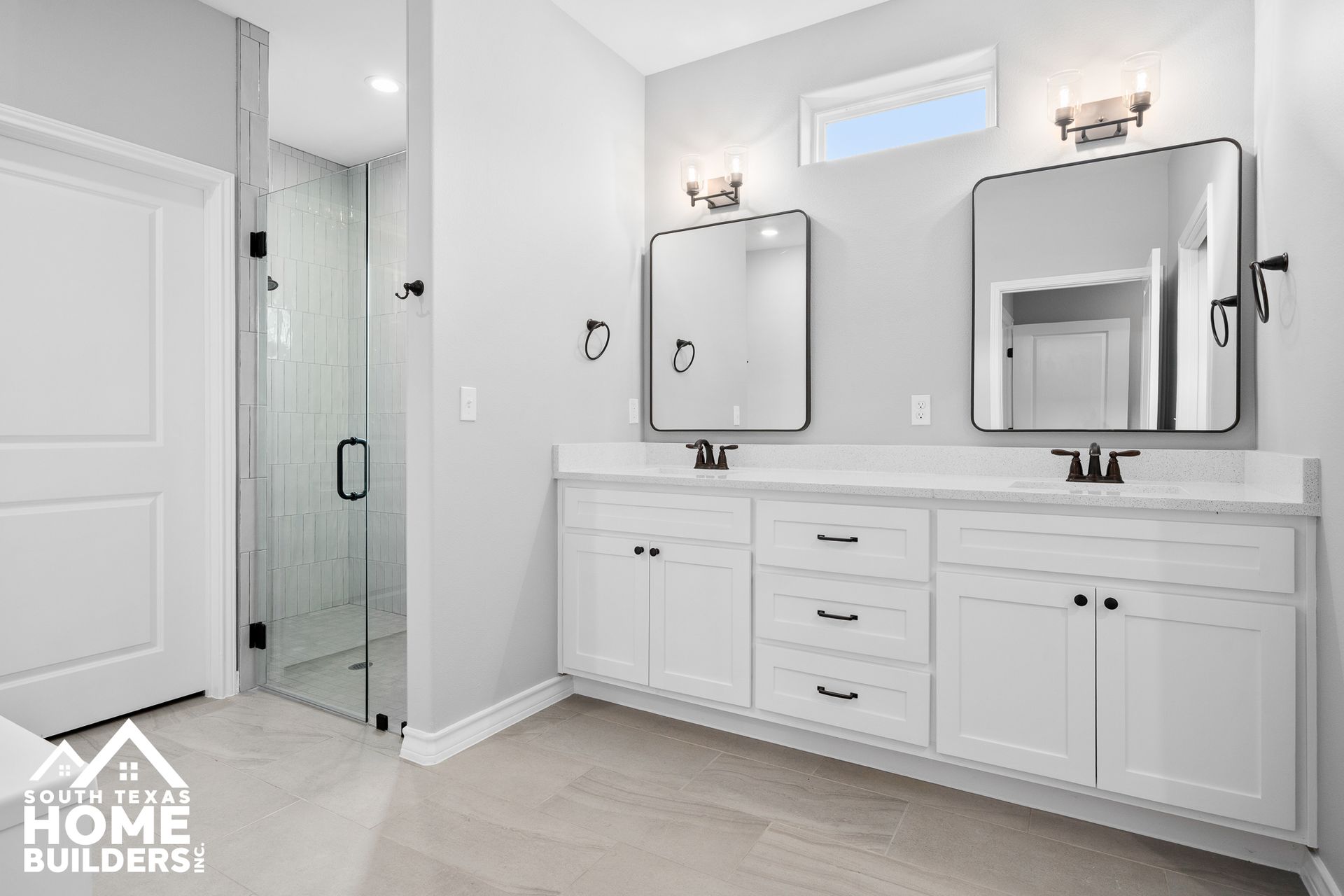 Spacious, modern bathroom with double vanity, mirrors, and glass shower. White cabinets and light gray walls.