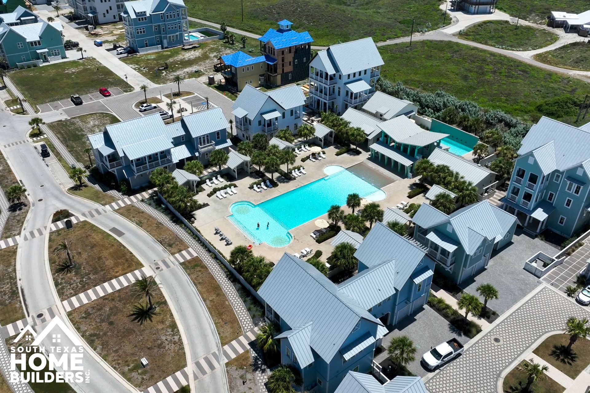 Aerial view of a coastal resort with buildings, pool, and surrounding green areas, all in a sunny outdoor setting.