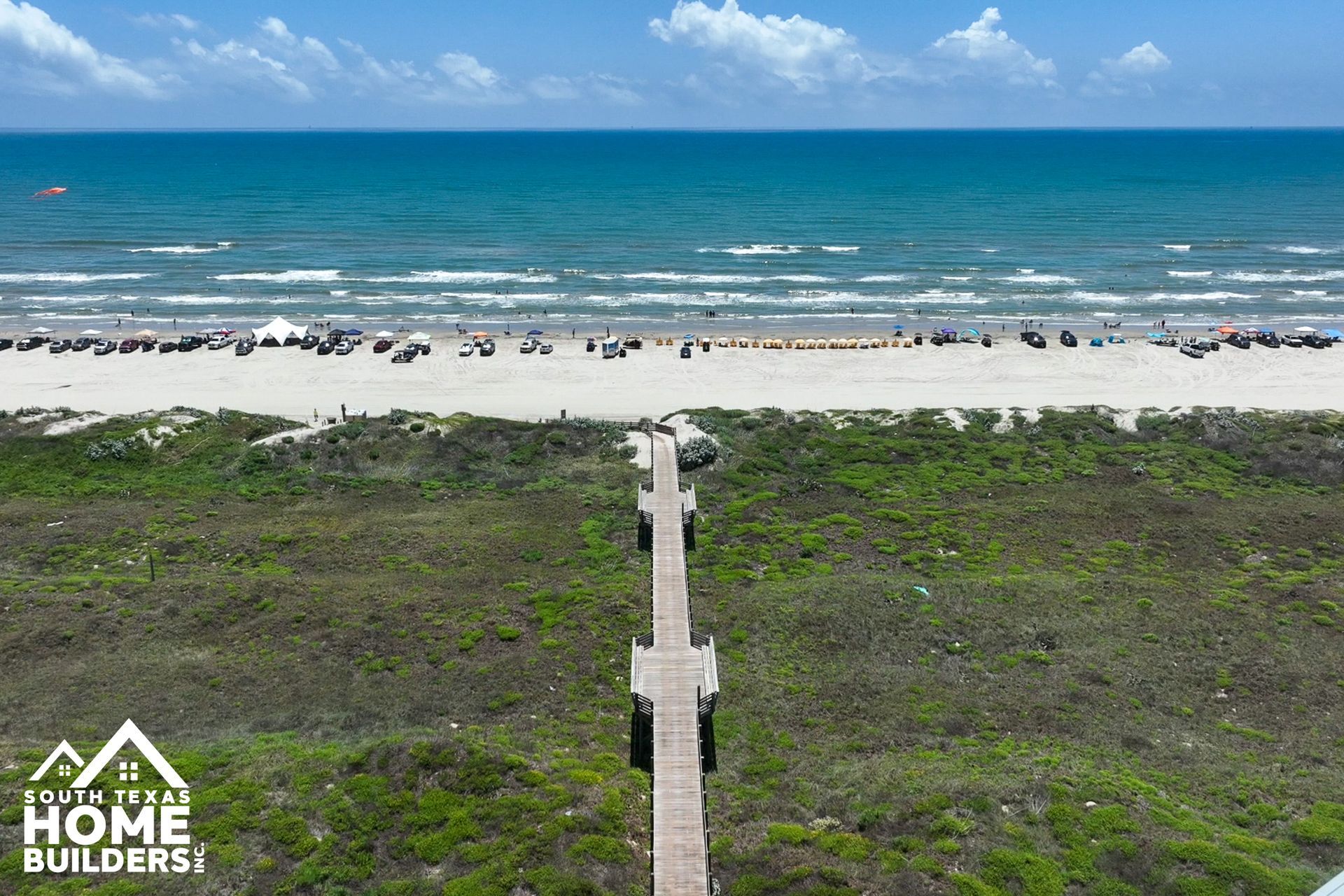 A boardwalk path leads to a crowded beach and ocean under a blue sky.