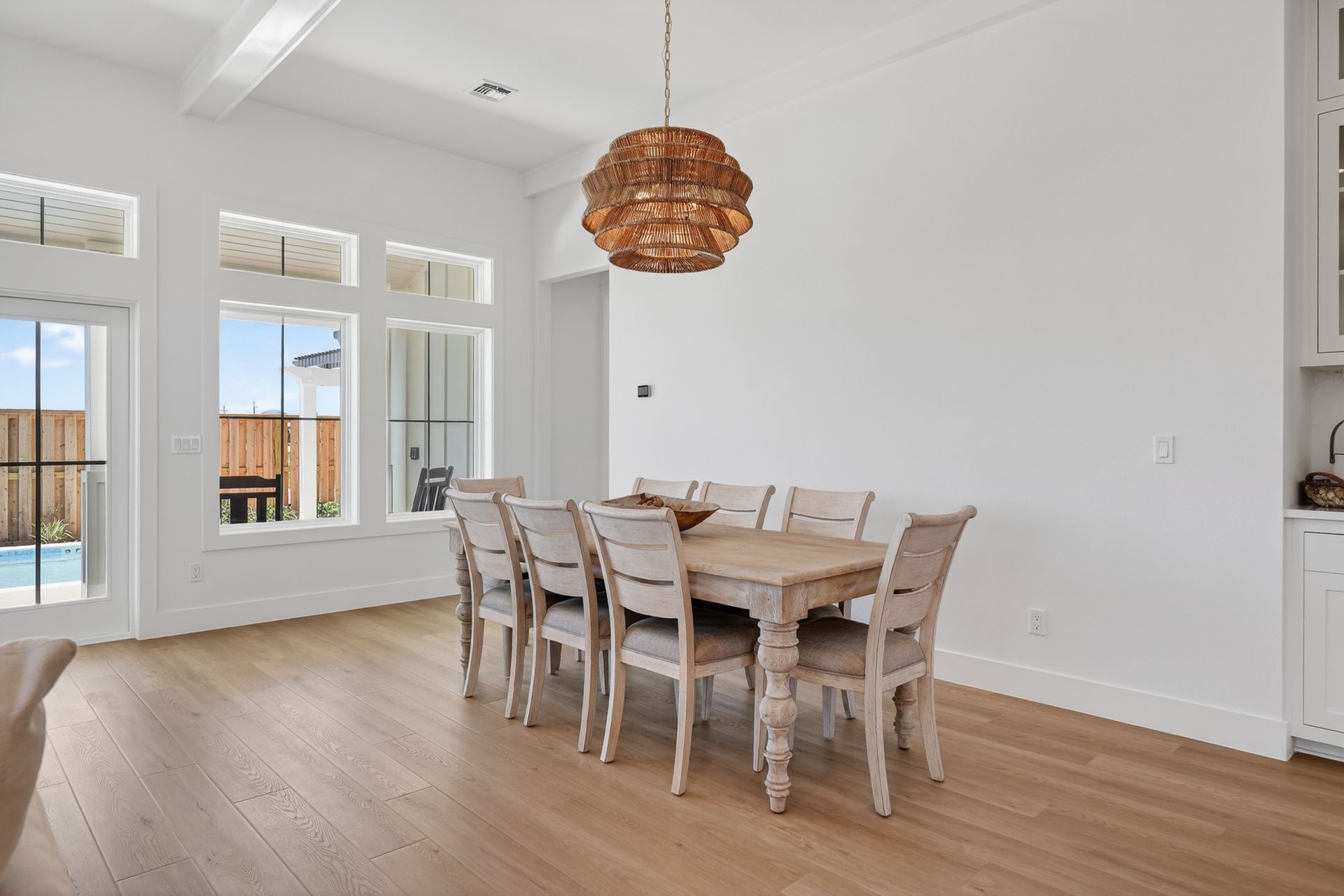 Bright dining room with a wooden table, chairs, woven pendant light, and large windows on a light wood floor.