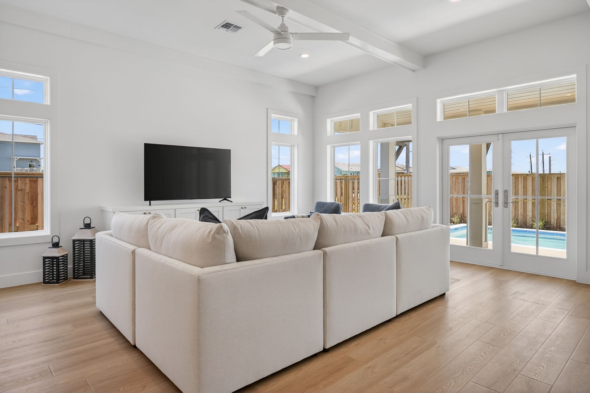 Bright living room with beige sectional sofa, wall-mounted TV, and large windows overlooking a pool