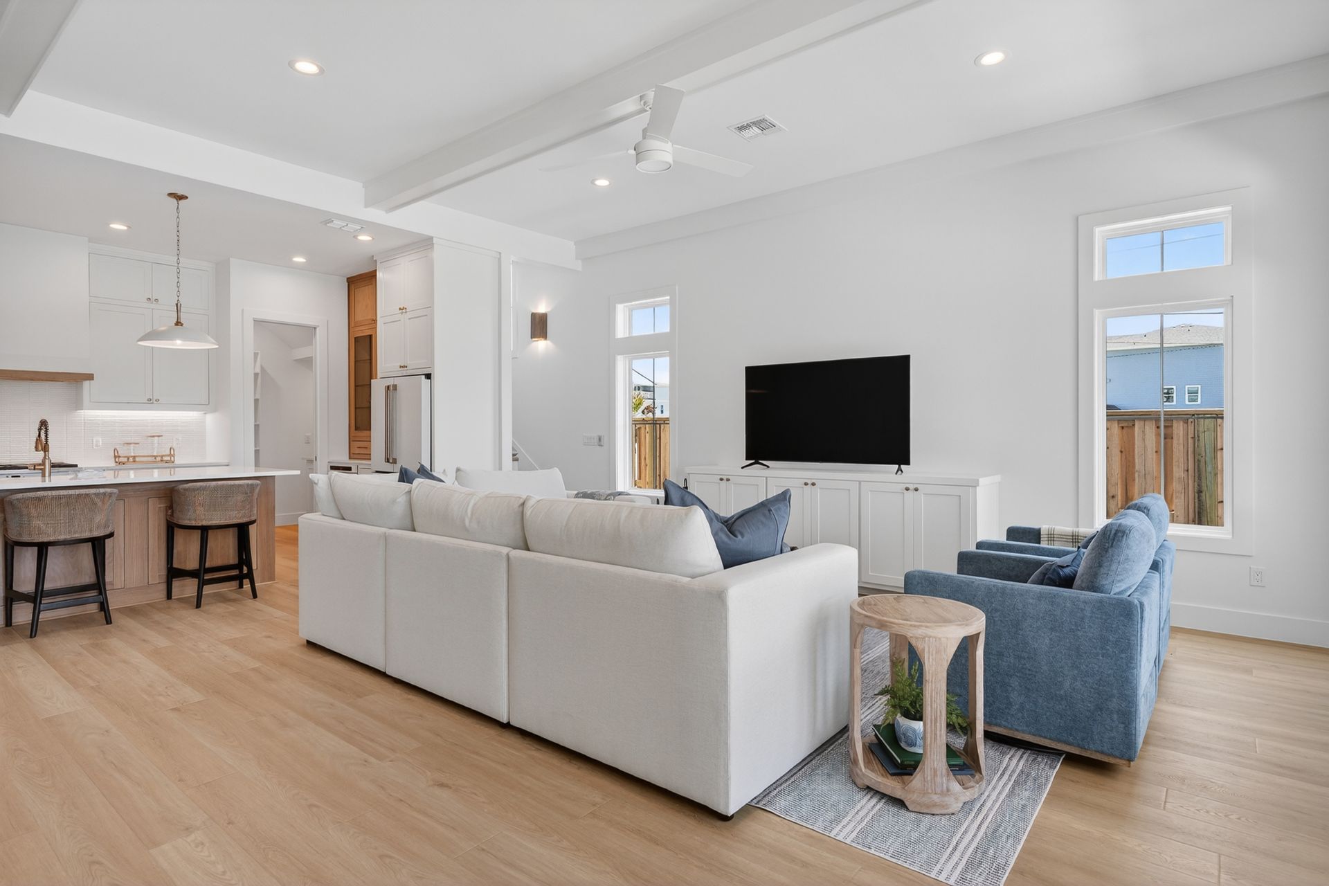 Bright open-plan living room with white sofa, blue chairs, wall TV, and kitchen island in the background