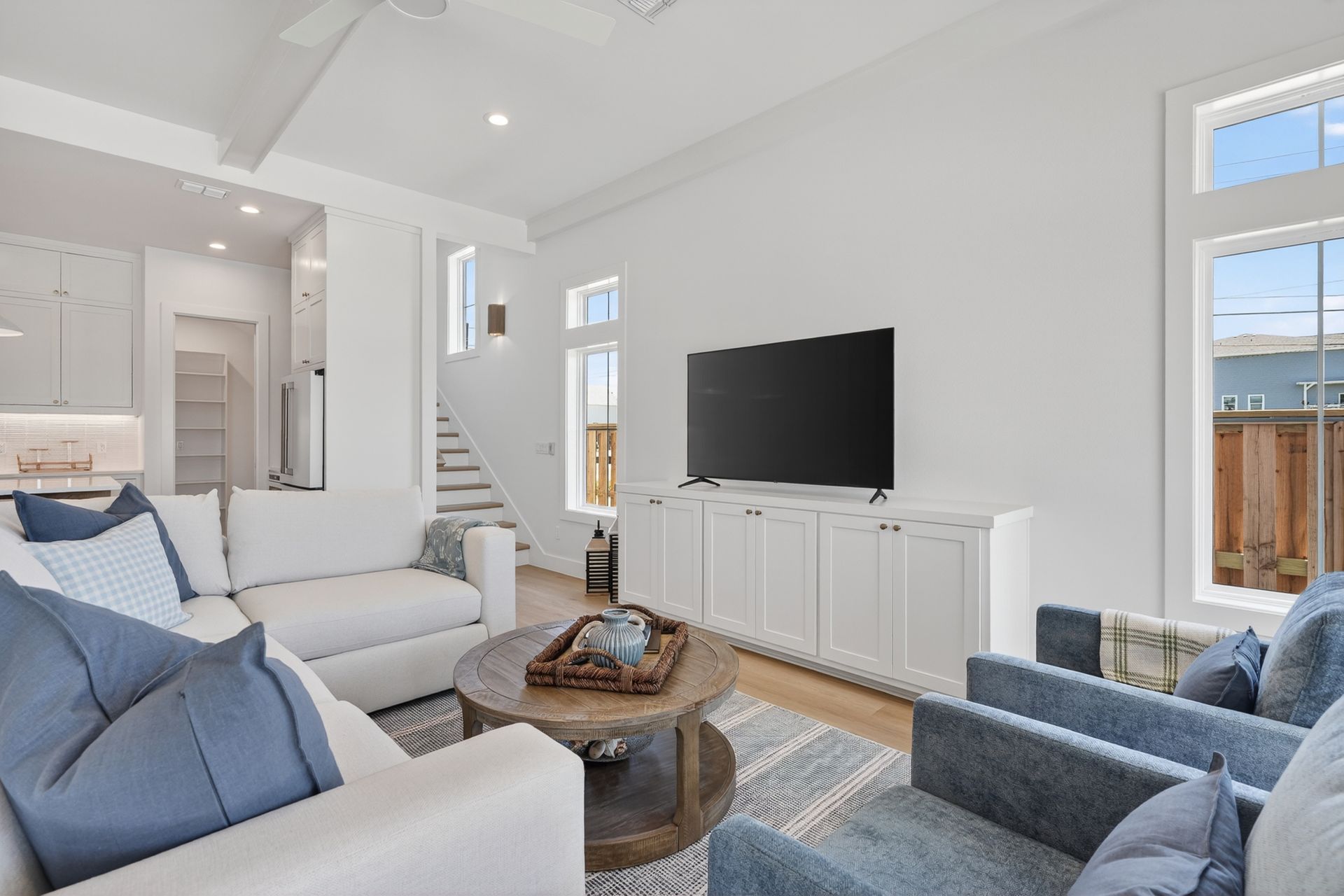 Bright modern living room with white sofas, blue chairs, a TV, and open kitchen in the background