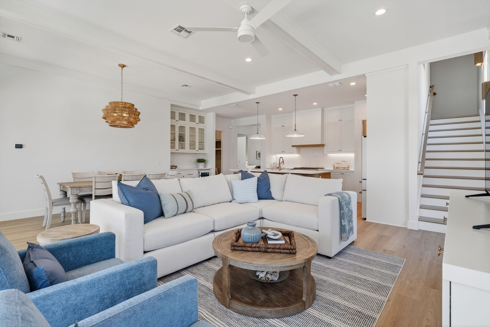Bright open-concept living room with white sectional, blue chairs, wood coffee table, and kitchen beyond