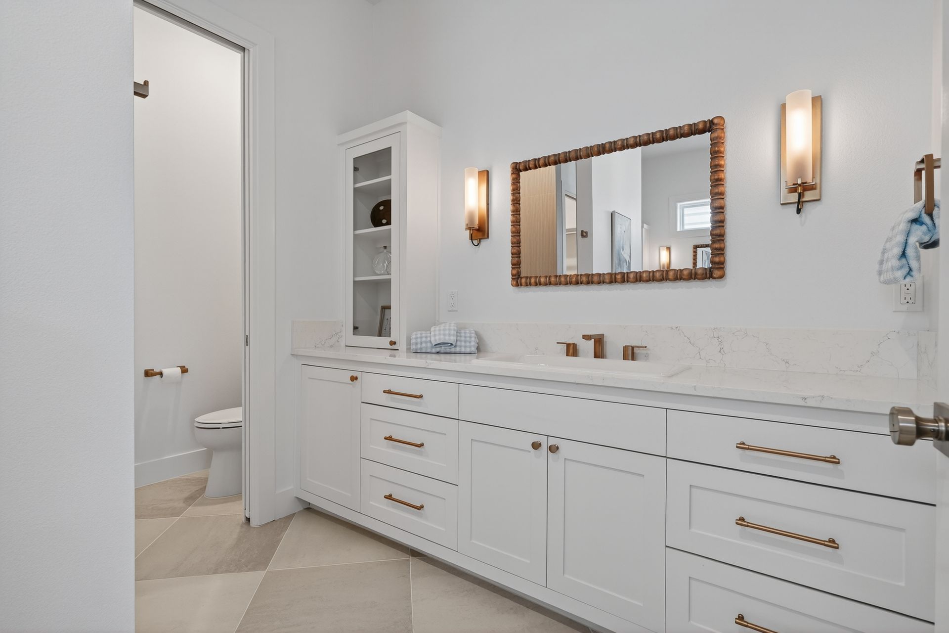 Bright white bathroom with double vanity, wall sconces, mirror, and open door to toilet room.