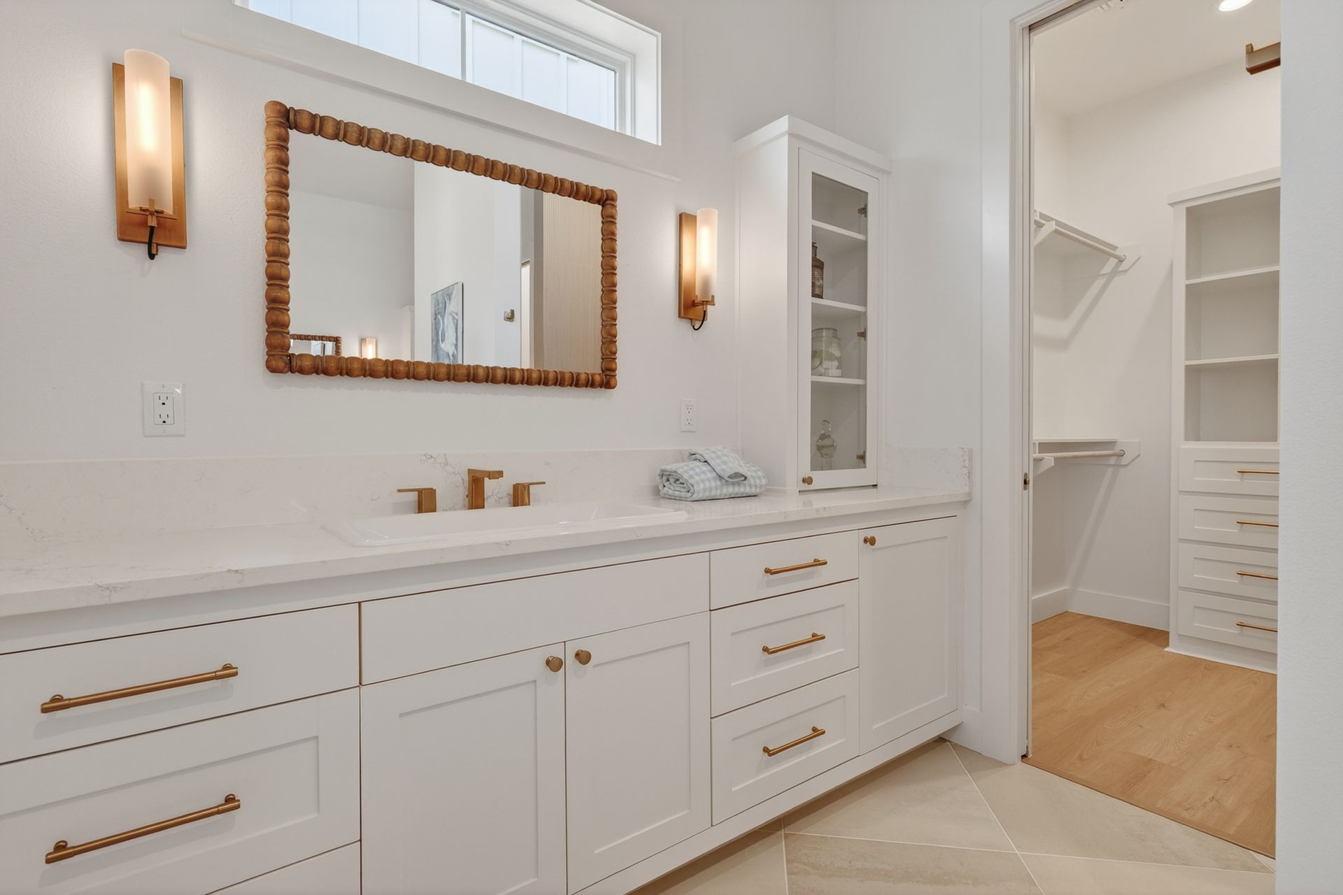 Bright white bathroom vanity with gold accents, mirror, sconces, and open doorway to a walk-in closet.