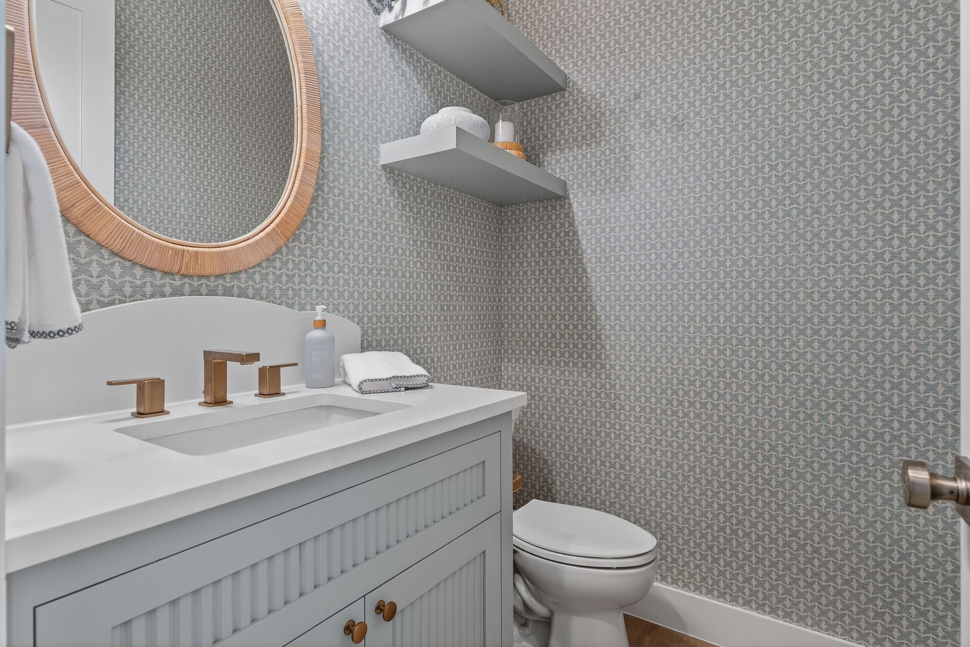 Small modern bathroom with patterned gray wallpaper, white vanity, round mirror, toilet, and wall shelves.
