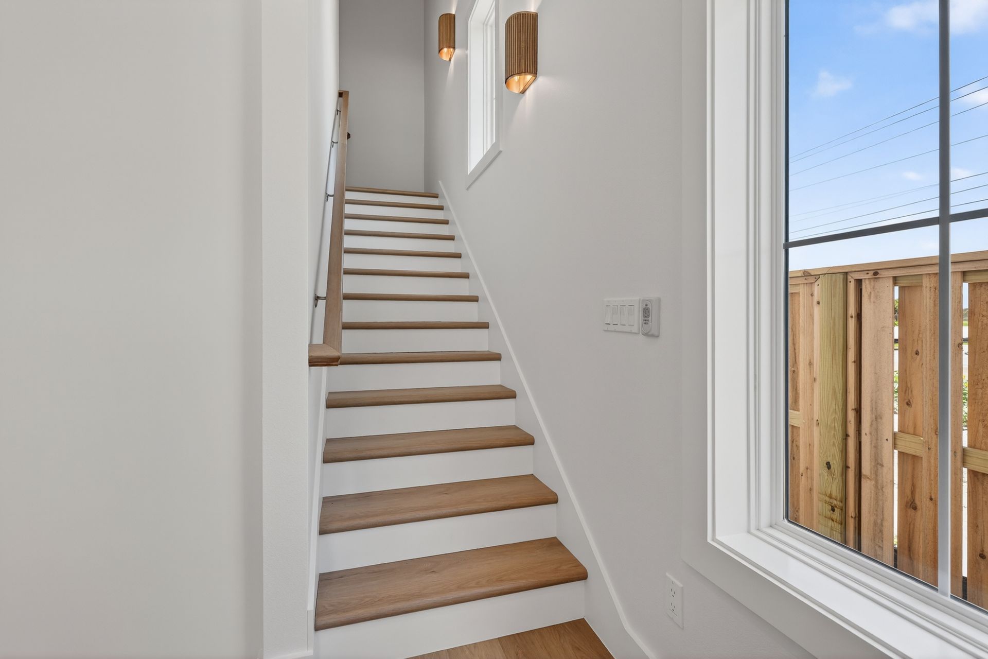 Narrow staircase with wood steps and white walls beside a bright window overlooking a fenced yard