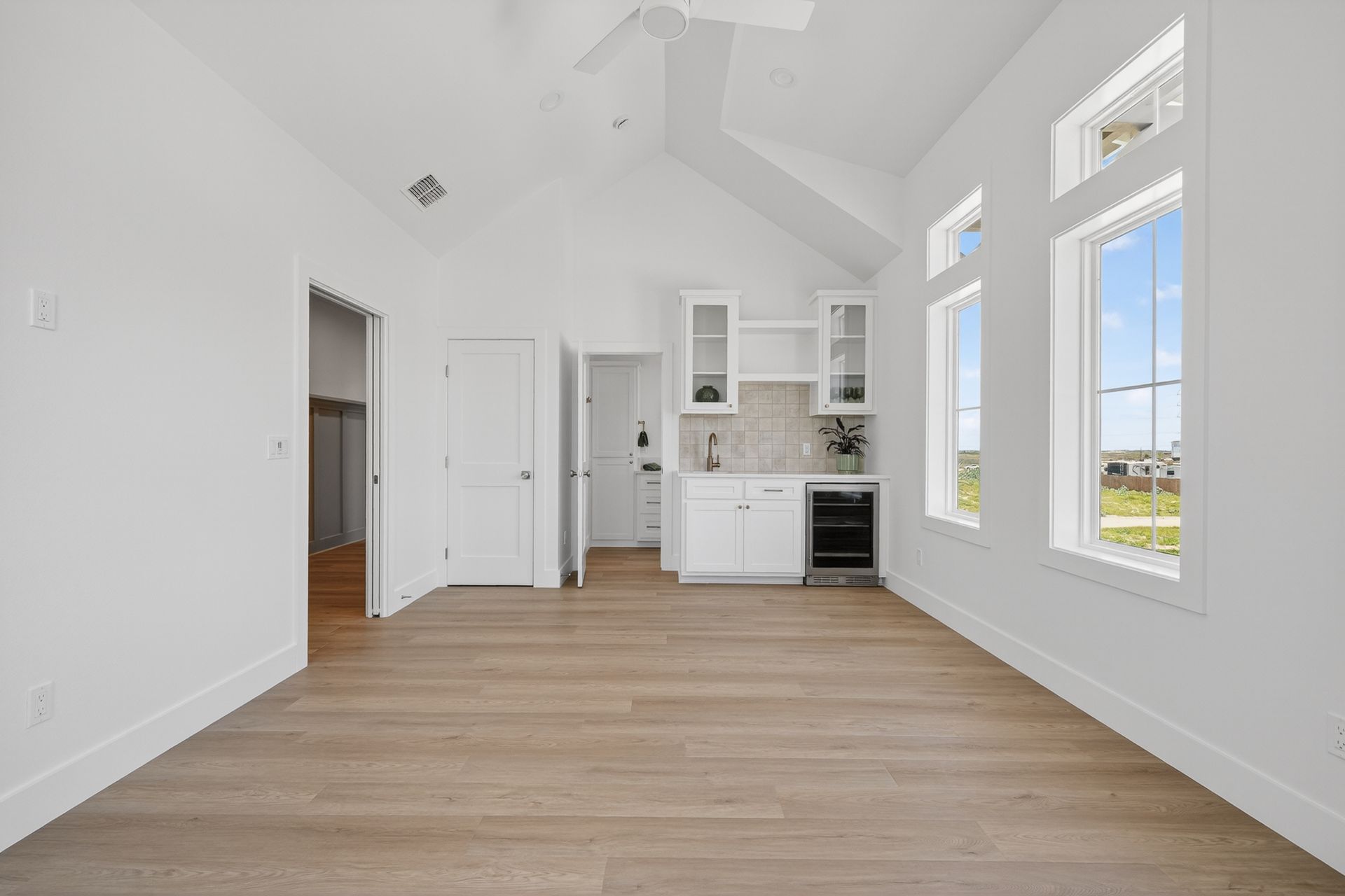 Open living room with vaulted ceiling, wood floors, white walls, kitchen alcove, and tall windows with countryside view