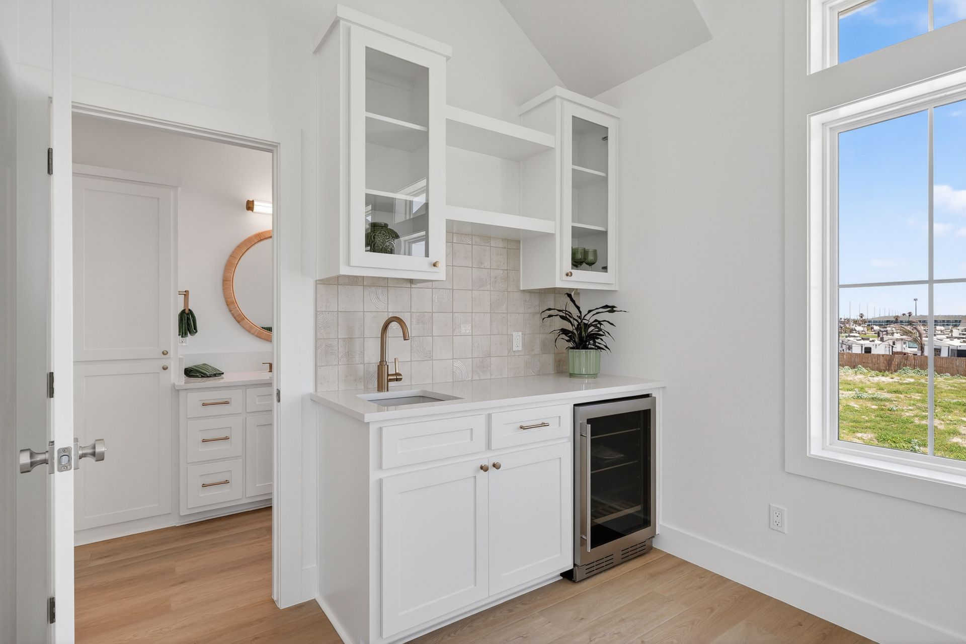 Bright white kitchen with open cabinets, sink, wine cooler, and large windows overlooking a yard.
