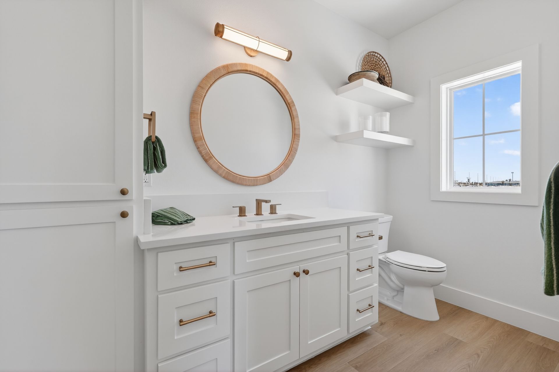 Bright white bathroom with wood vanity, round mirror, floating shelves, and a window