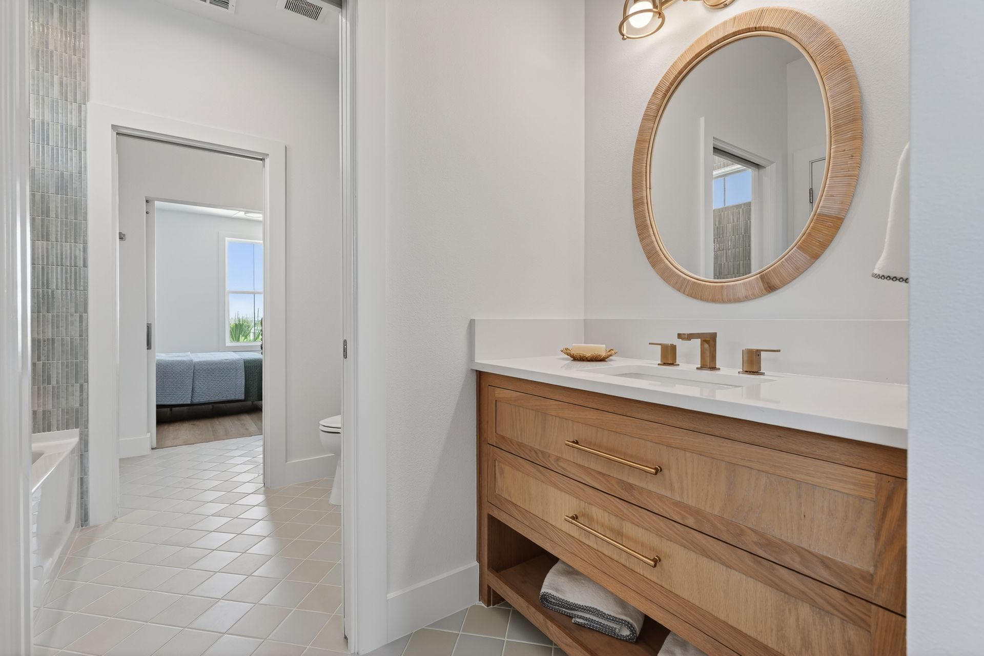Bright bathroom vanity with round gold mirror, white walls, and a doorway to a bedroom.