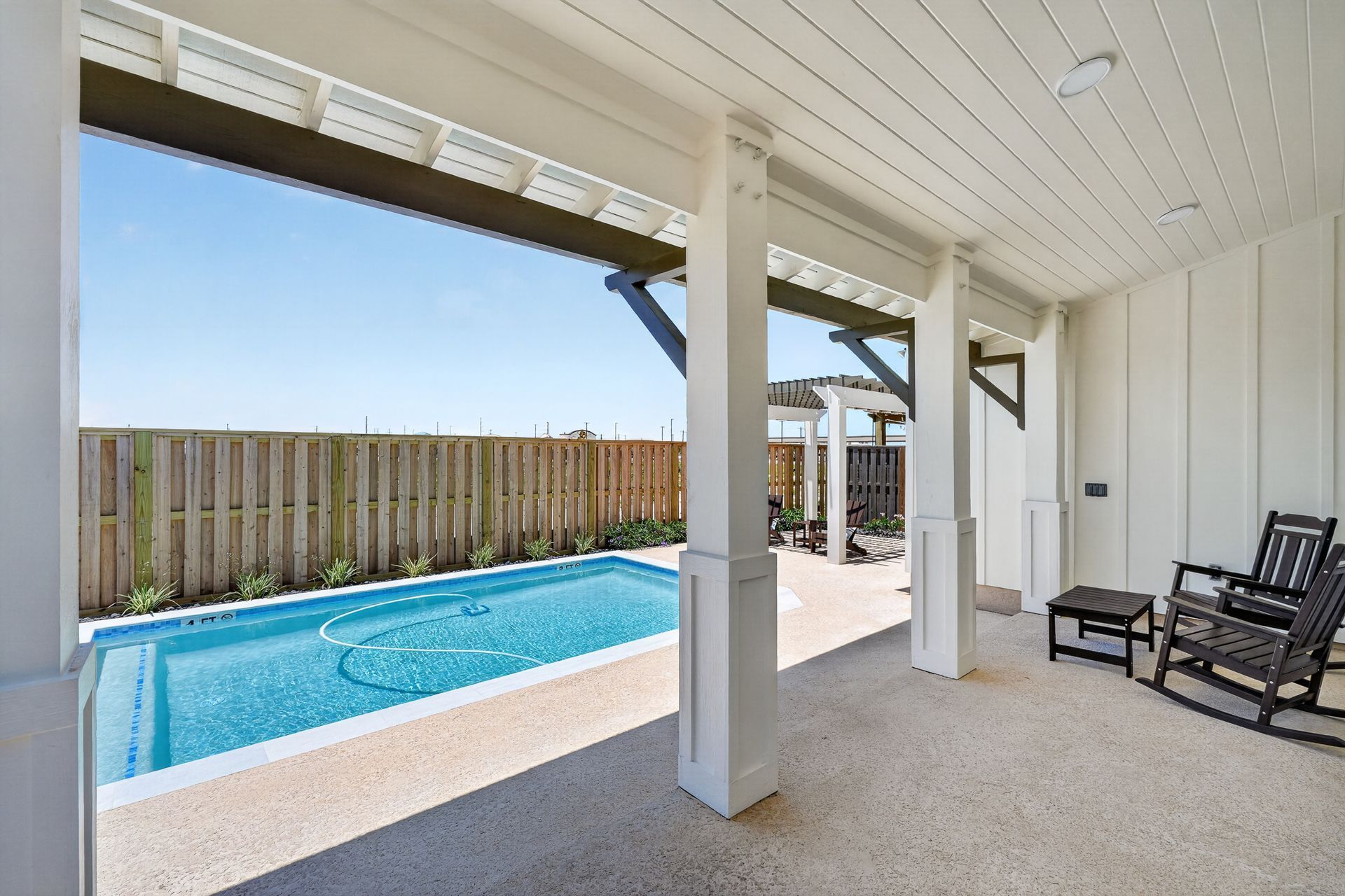 Covered patio beside a turquoise pool, with white posts, wood fence, and black lounge chairs.