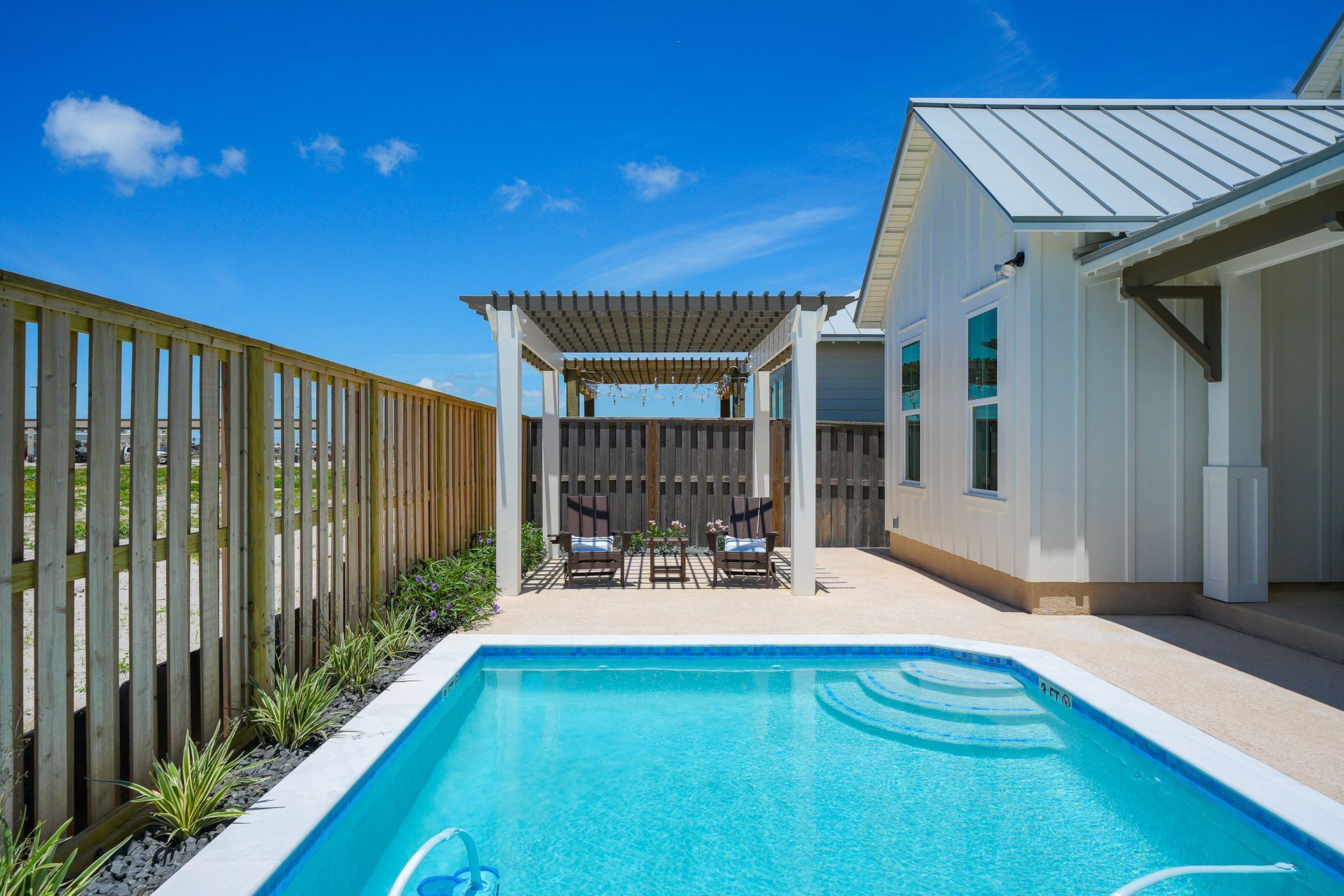Modern patio with a turquoise pool, wooden fence, pergola, and white house under a clear blue sky