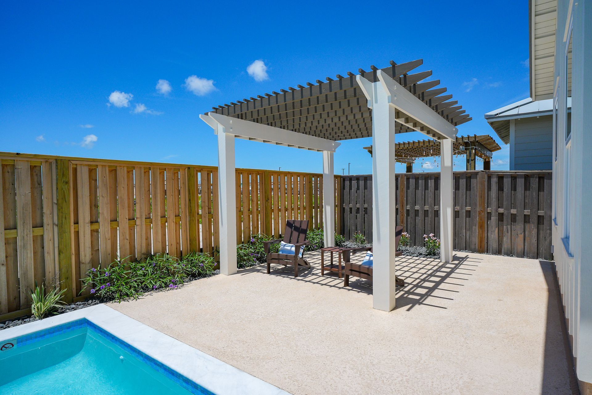 Backyard patio with white pergola, wooden fence, lounge chairs, and a swimming pool edge under a blue sky