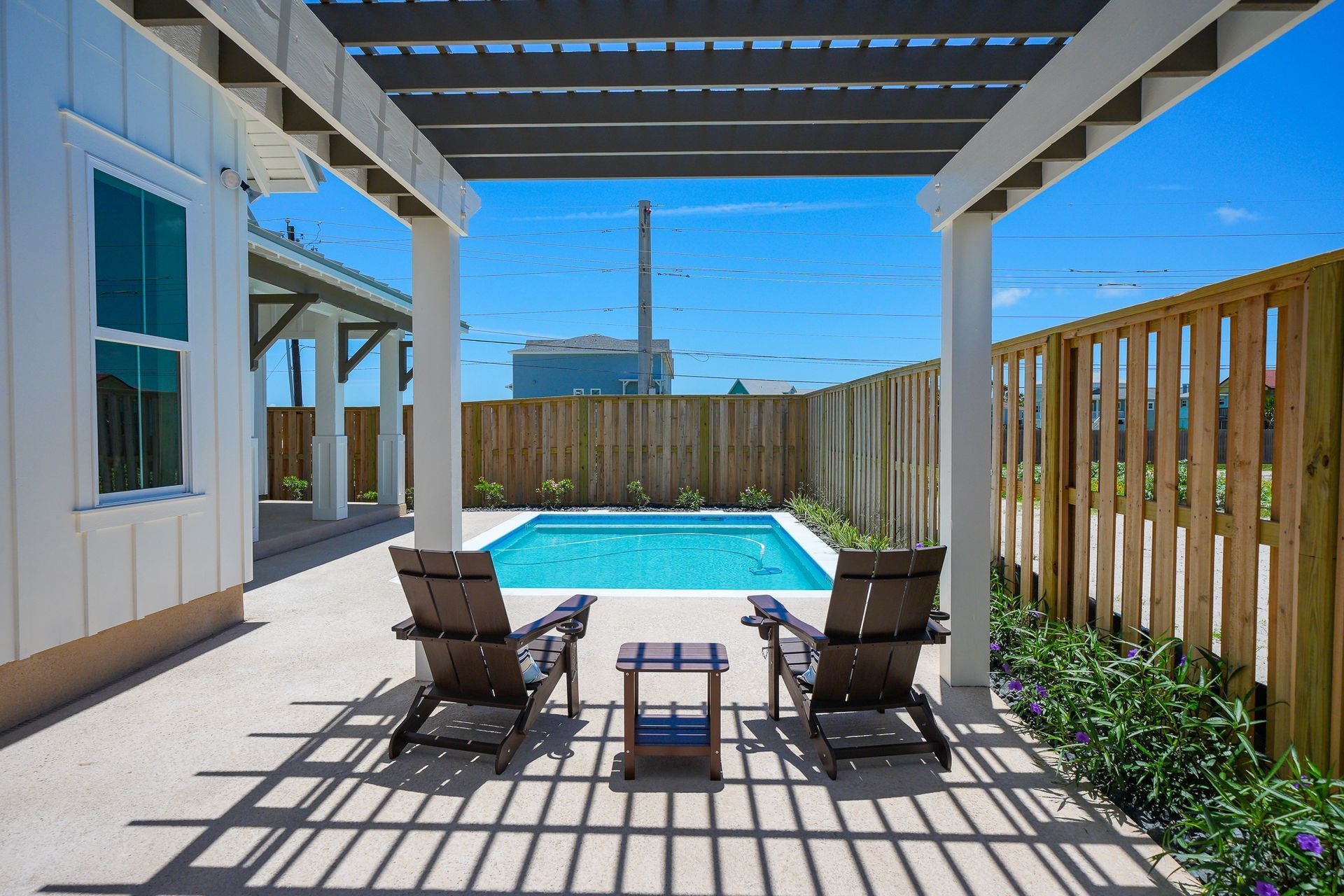 Covered patio with two chairs facing a backyard pool and tall wooden fence under a clear blue sky