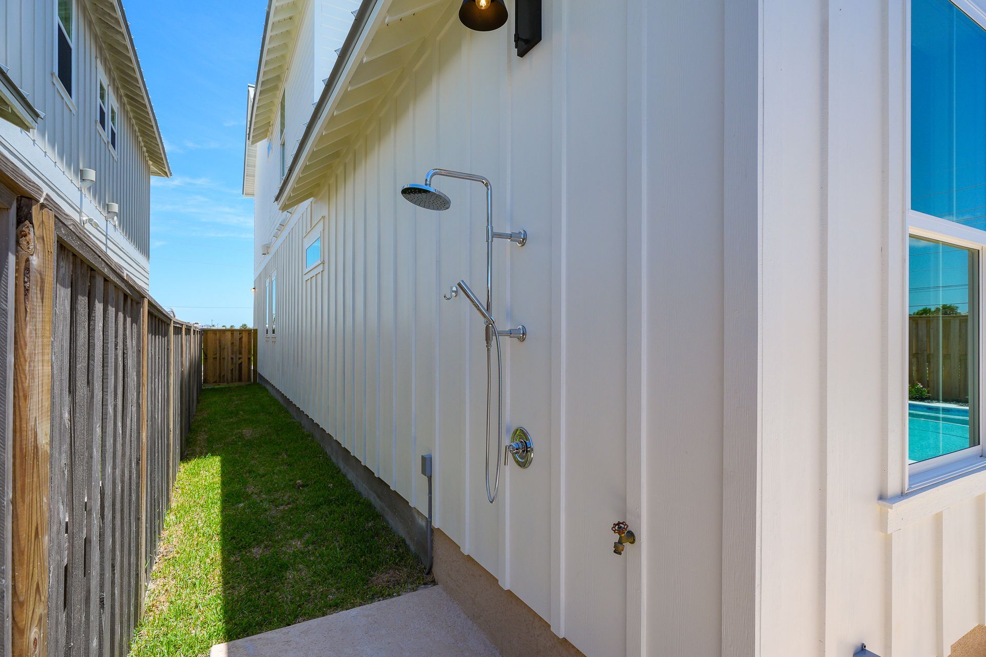 Narrow side yard beside a beige house with outdoor shower fixtures and a strip of grass along the fence.