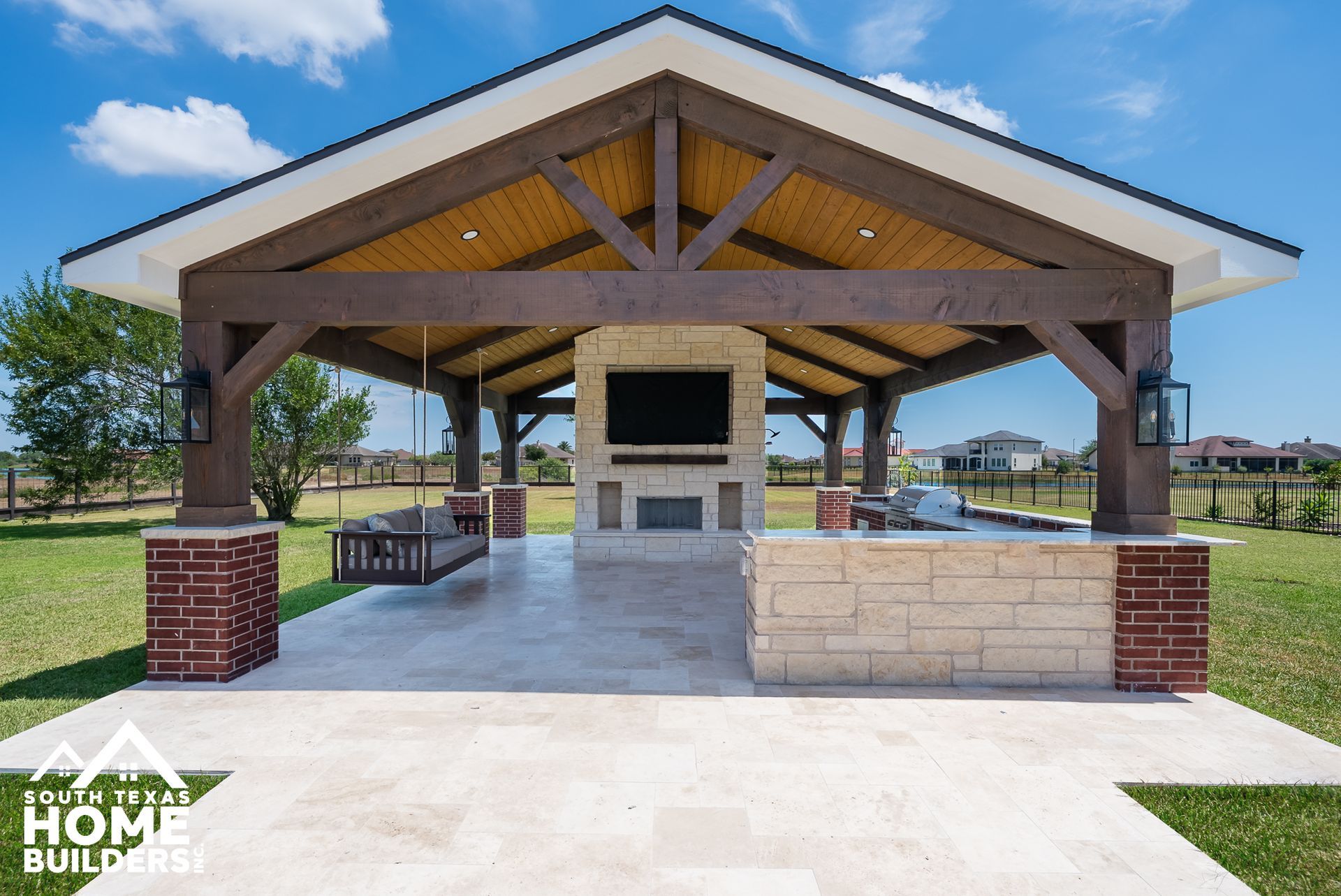 Outdoor pavilion with brown beams, a fireplace, and a television on a sunny day.