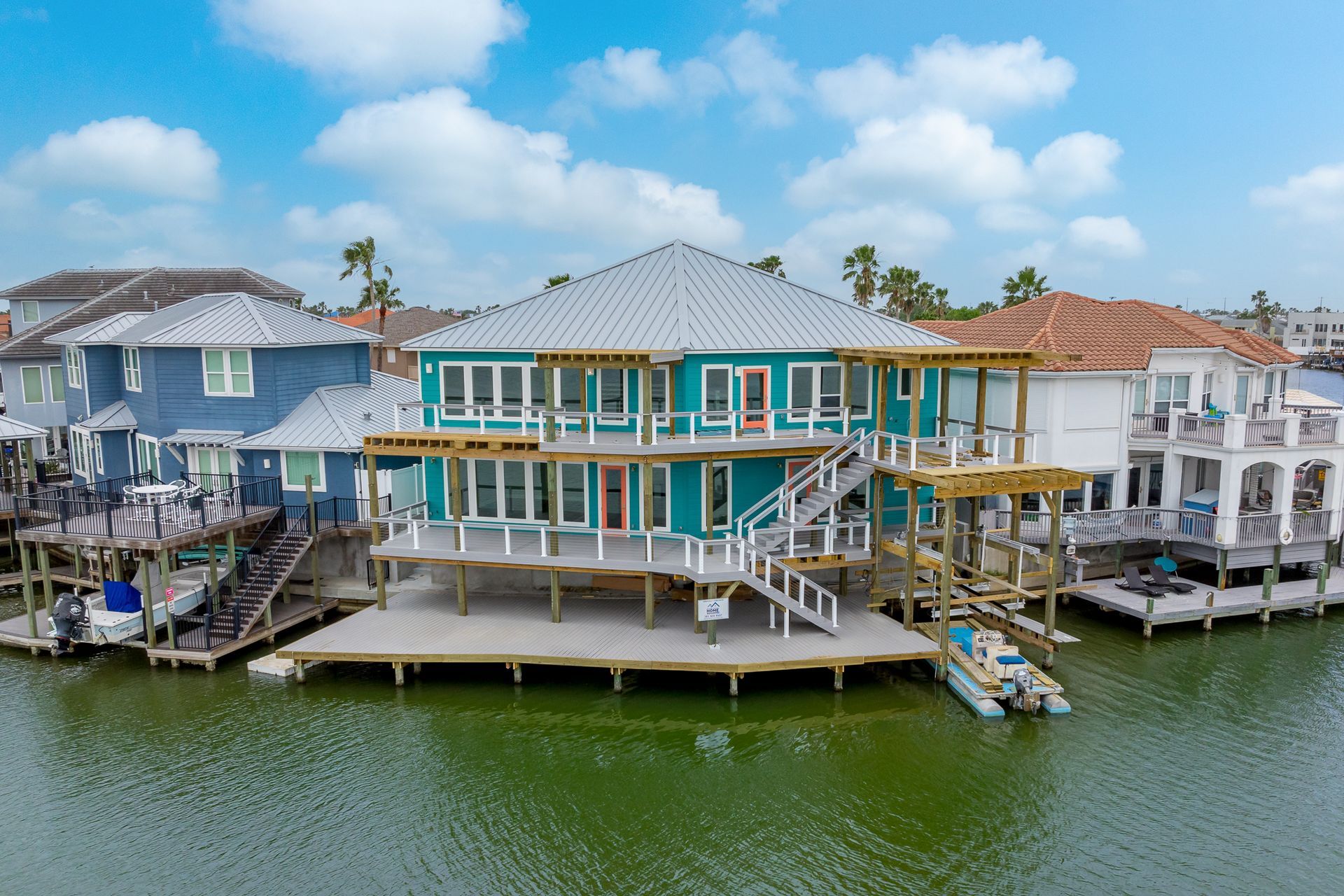 Two-story teal house on stilts with boat dock, on a canal, with neighboring houses.