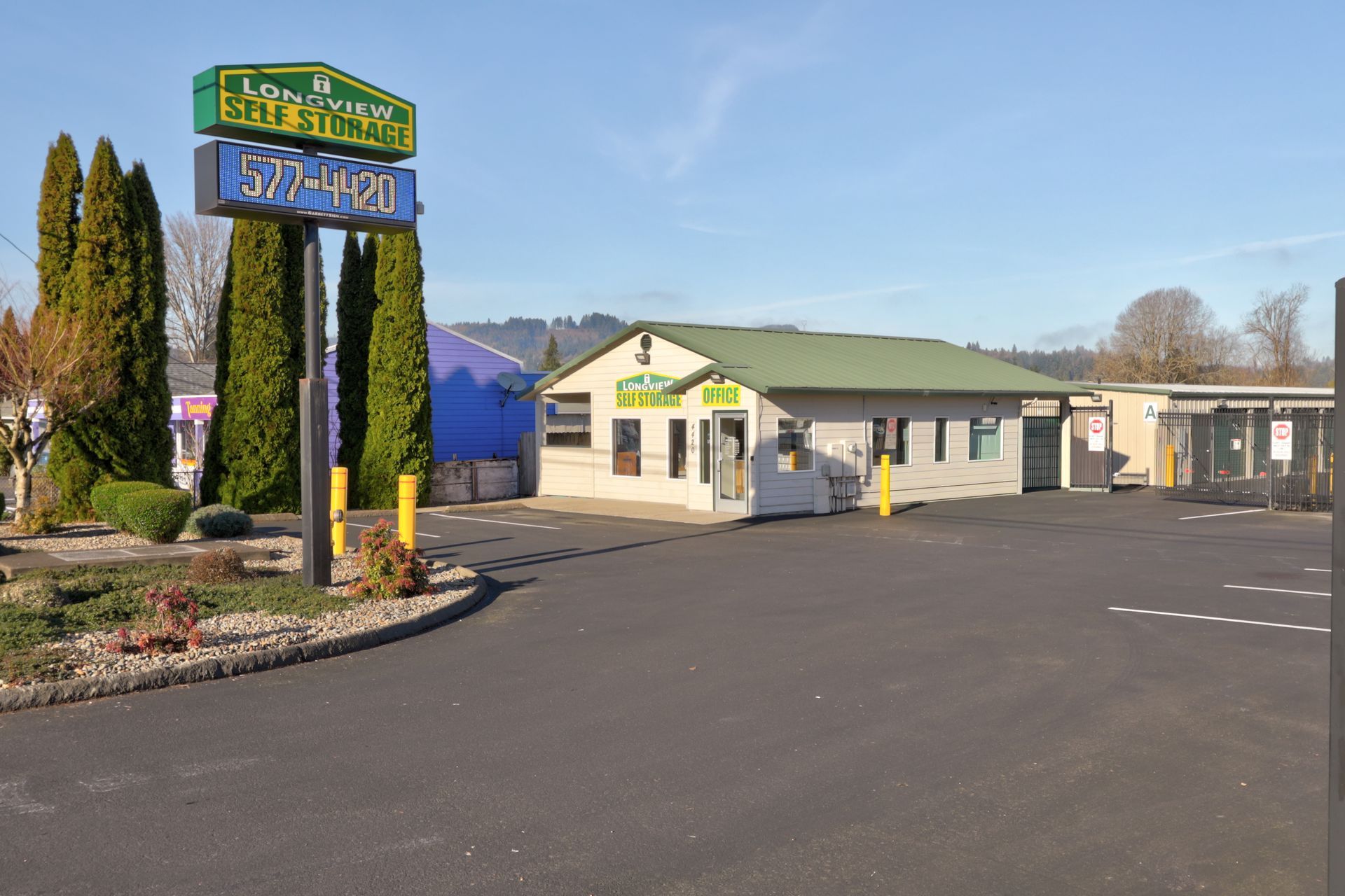 Storage business with sign, beige building, asphalt lot, green roof, and some trees.