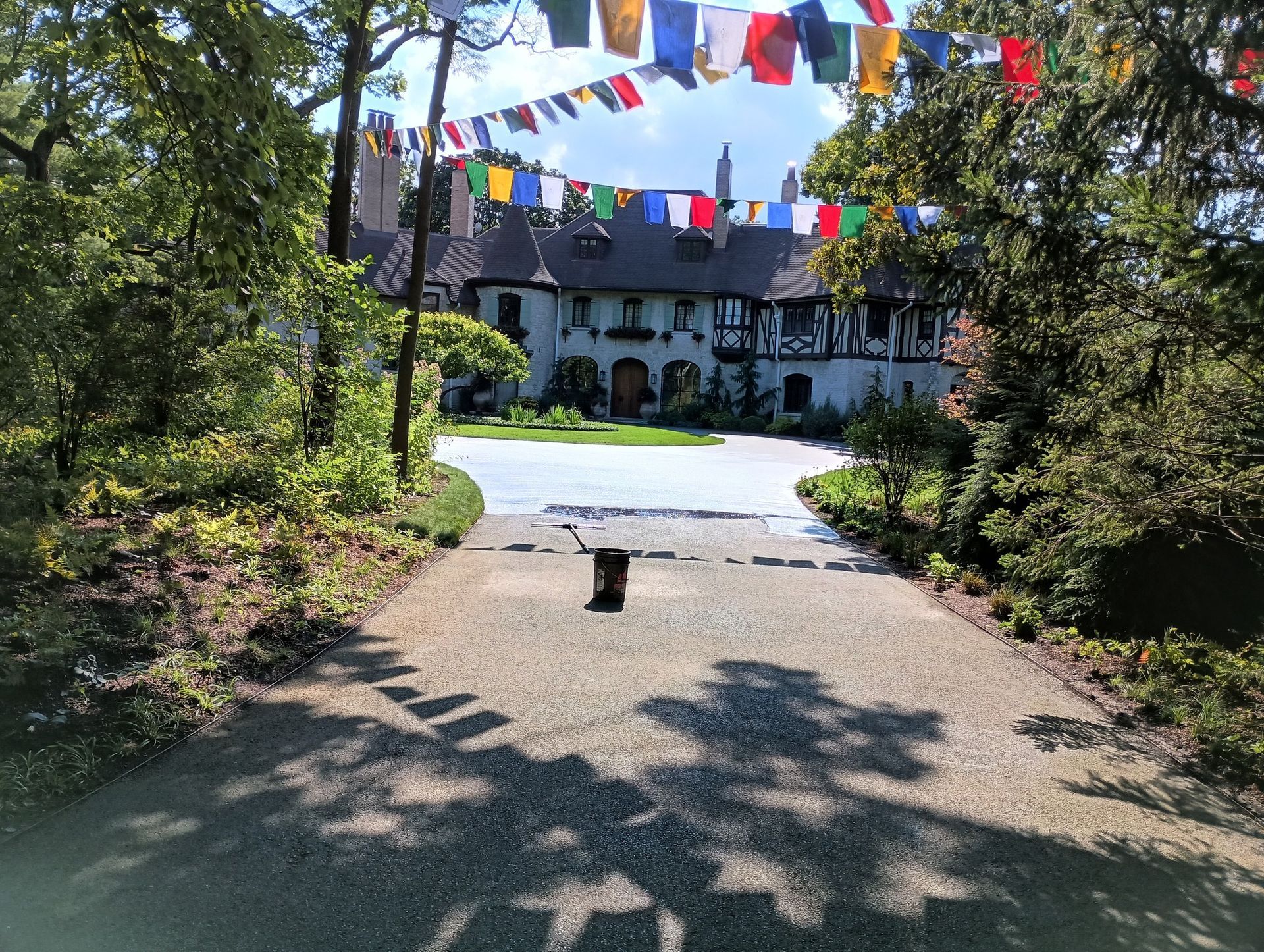 Gravel driveway leads to a Tudor-style house with colorful flags strung above. Trees frame the view.