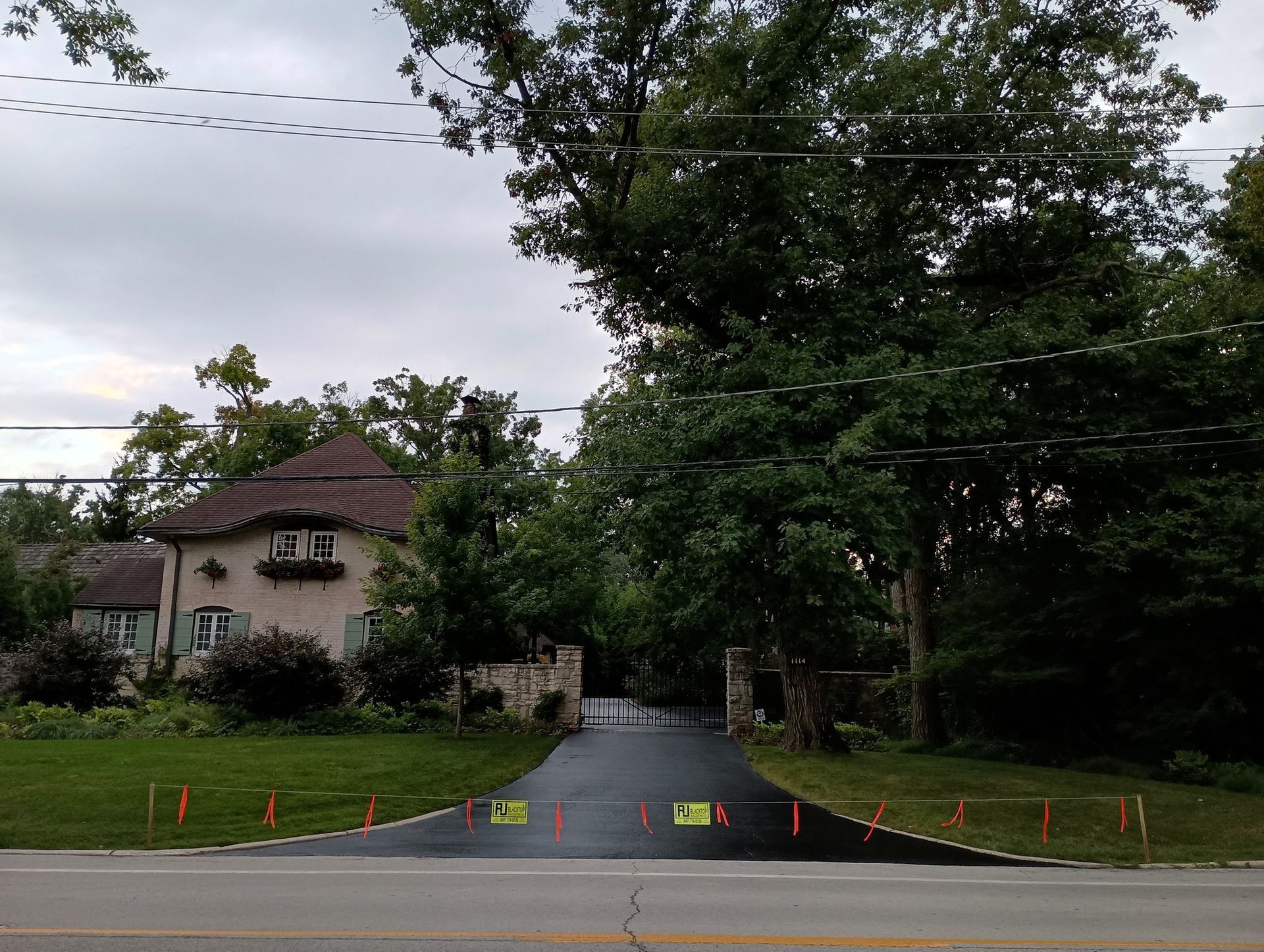 Driveway with fresh asphalt, lined with orange markers, leading to a house with a brown roof, surrounded by trees.