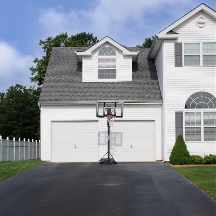 A two-story white house with a basketball hoop in front of the garage on a black driveway.