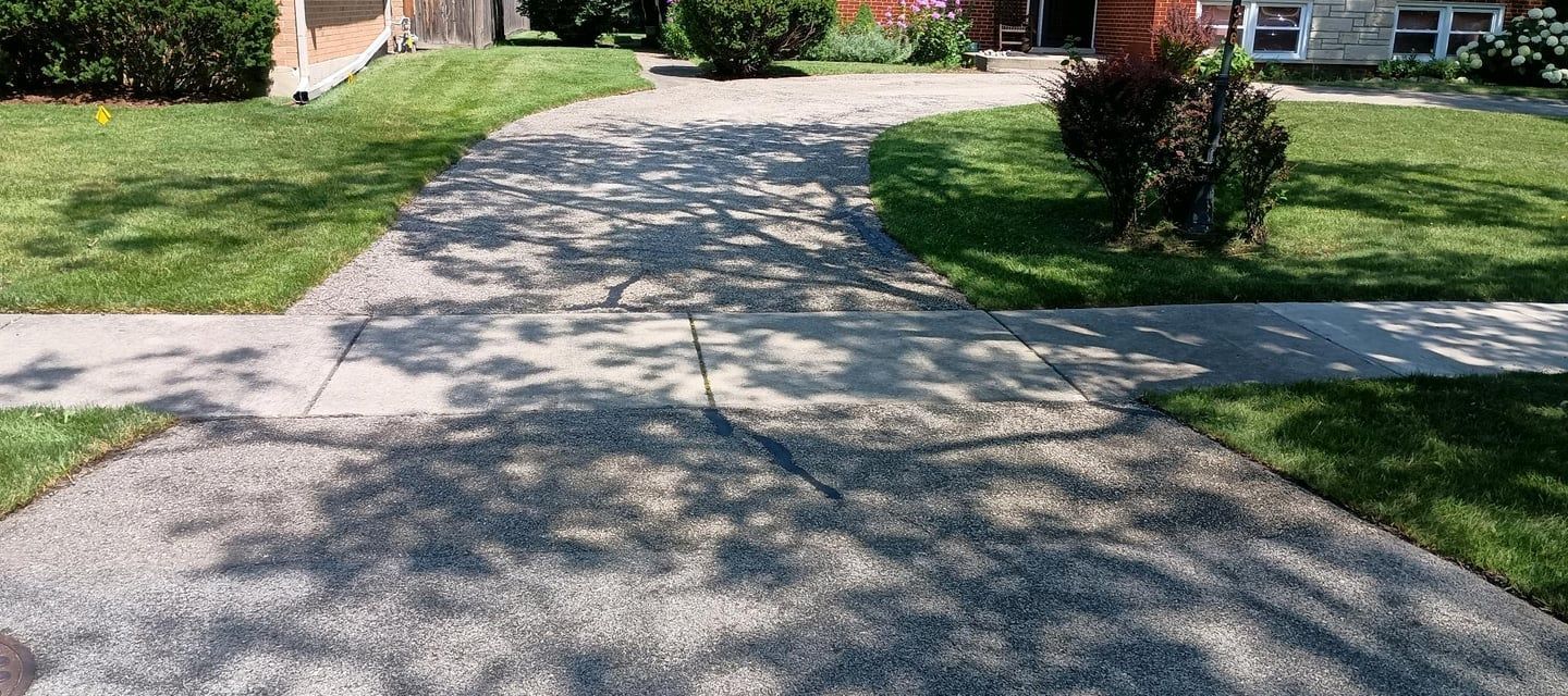 A cracked concrete driveway with tree shadows and green lawn in a sunny residential area.