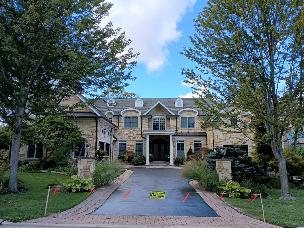 Newly paved black driveway leading to a blue-gray house with a white garage door, on a sunny day.
