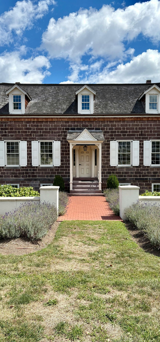 A brown shingled house with a white front door and trim, flanked by white shutters, accessed by a red brick walkway.