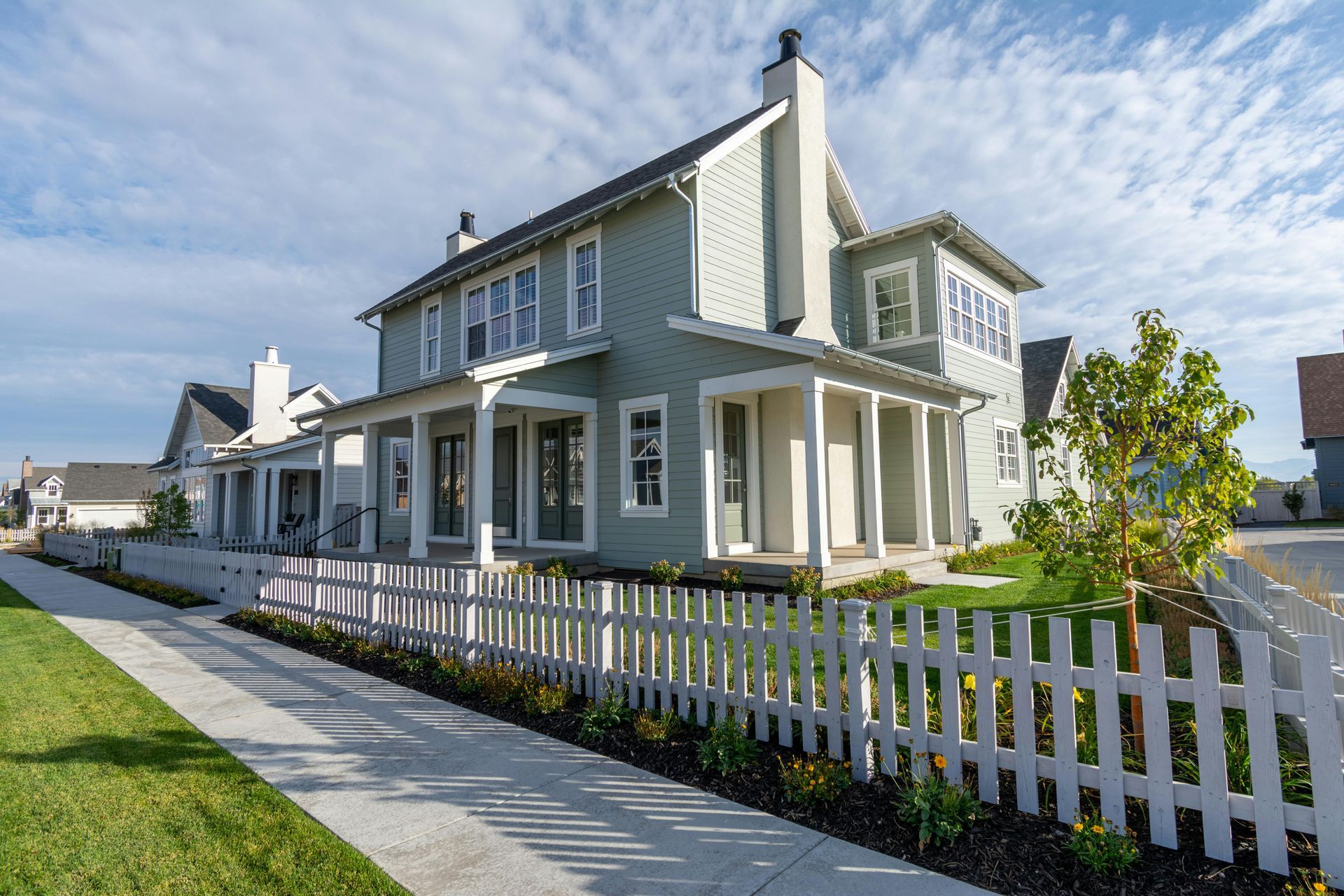 A light green two-story house with a white picket fence, covered porch, and blue sky in a quiet residential neighborhood.