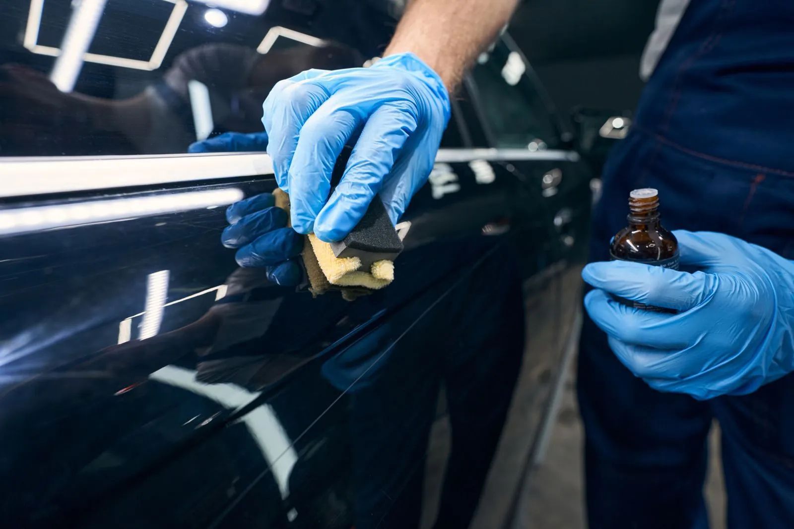 Blue-gloved hands applying polish to a black car door in a detailing shop
