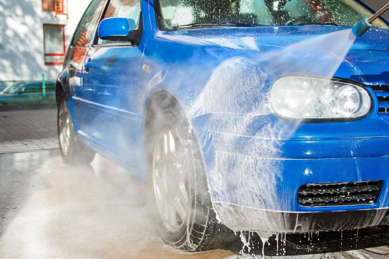 Blue sports car being washed with a high-pressure water spray, splashing across the front wheel and bumper.