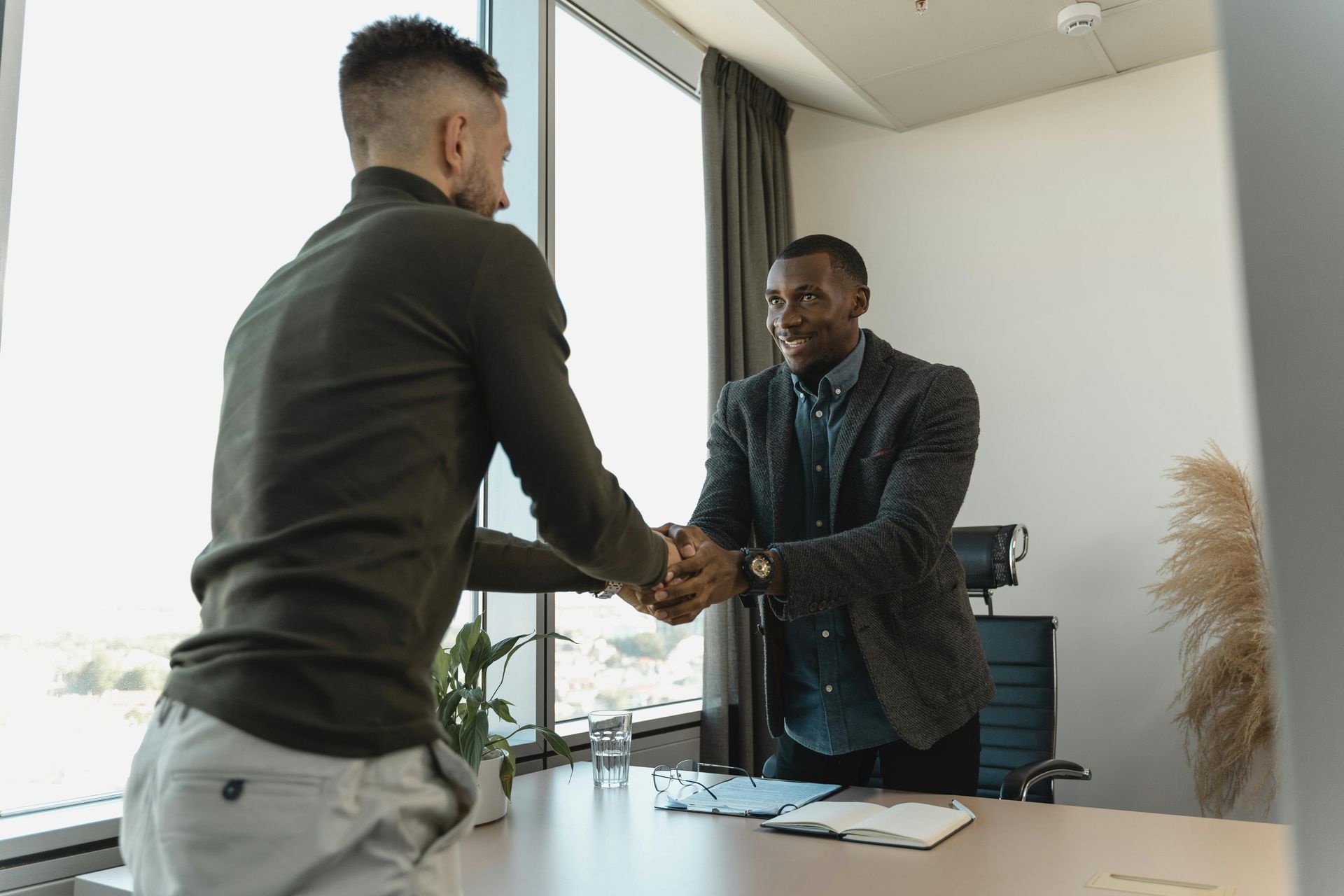 Two men are shaking hands in an office while sitting at a desk.
